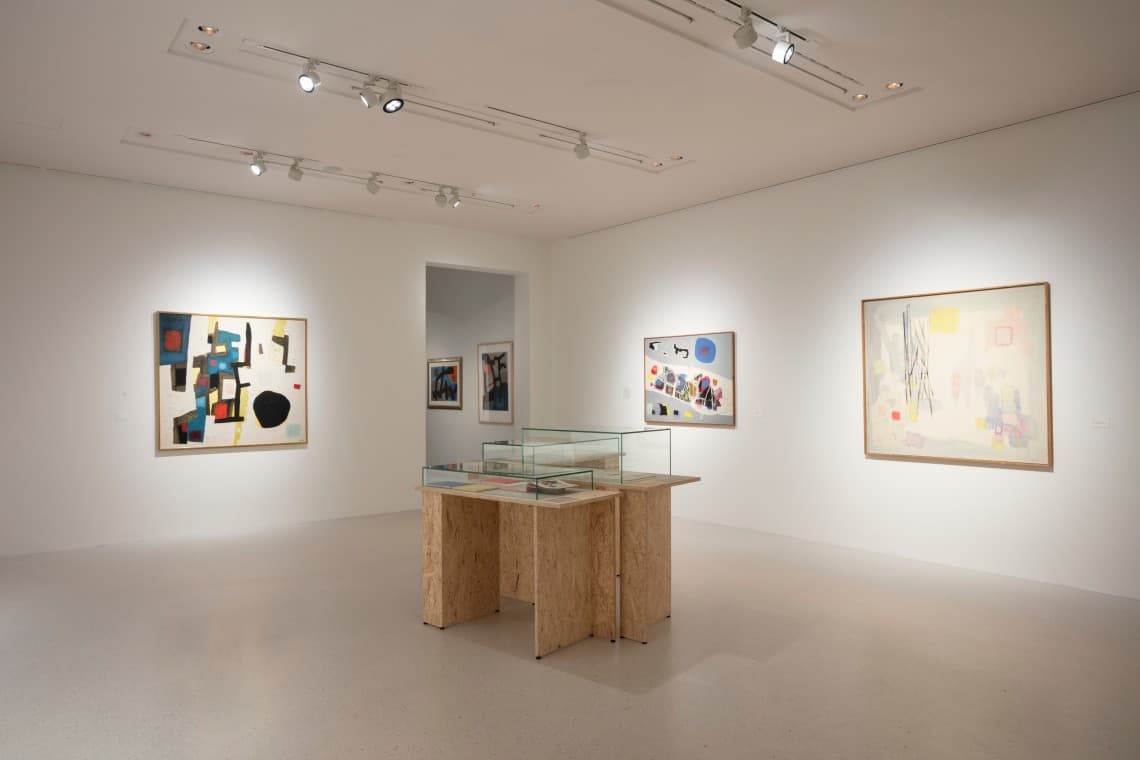 Modern art gallery with abstract paintings on white walls, a central glass display case, and overhead lighting.