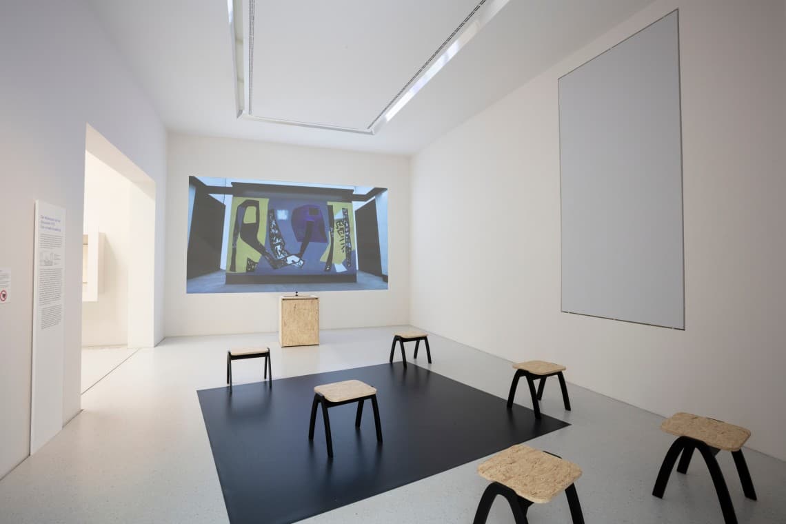 Modern art gallery room with a projection on the wall, five wooden stools on a black square mat, and a minimalist design.