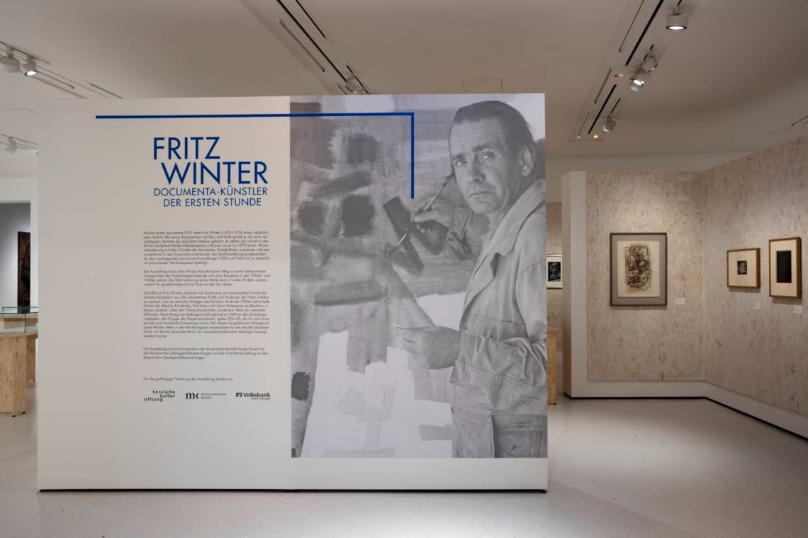 Exhibition display featuring a large panel with text and a black-and-white photo of at artist painting, surrounded by framed artworks.