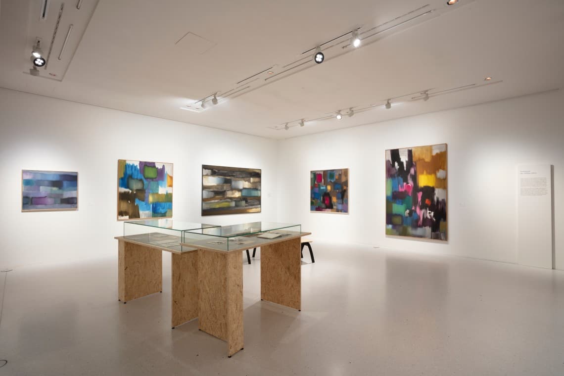 Art gallery with abstract paintings on white walls, glass display tables in the center, and overhead lighting.