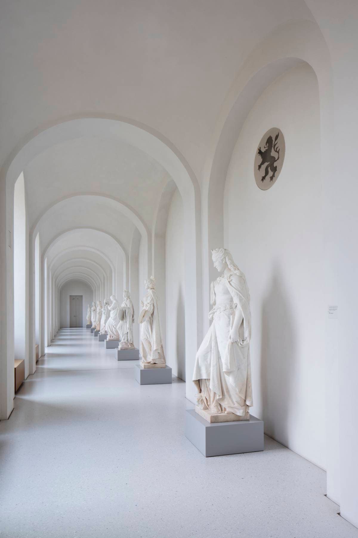 Ein langer, weißer Korridor mit gewölbten Decken und einer Reihe klassischer Statuen auf Sockeln an einer Seite.