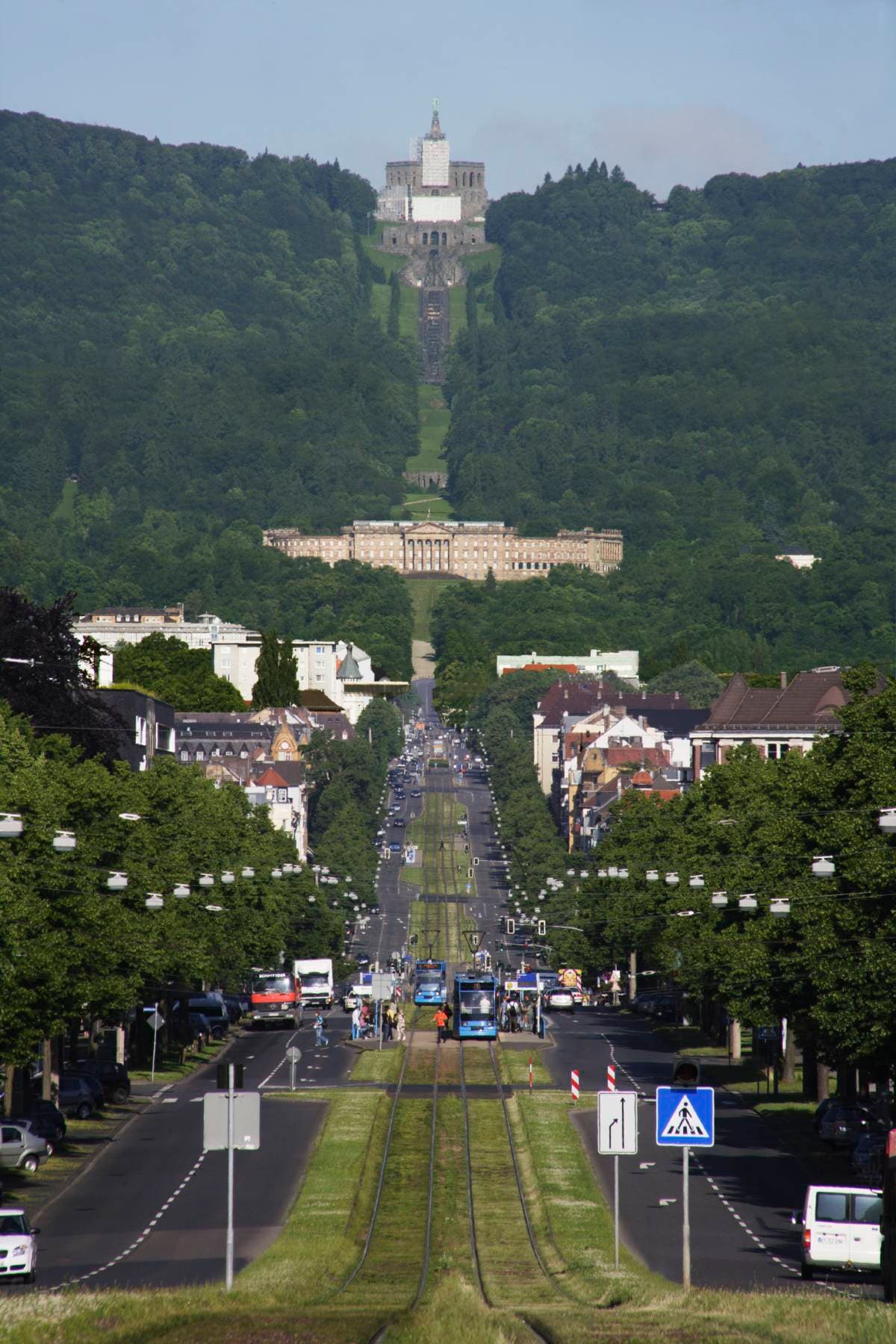 Blick auf eine lange, gerade Straße, gesäumt von Bäumen und Gebäuden, die zu einem Denkmal auf einem Hügel in einer üppigen, grünen Landschaft führt.