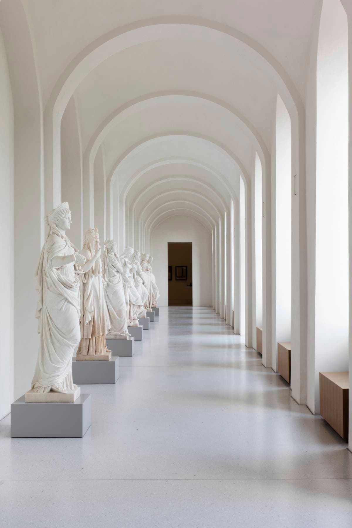 Eine Reihe weißer Marmorstatuen in einem langen, gewölbten Flur mit großen Fenstern auf der rechten Seite schaffen eine heitere und elegante Atmosphäre.