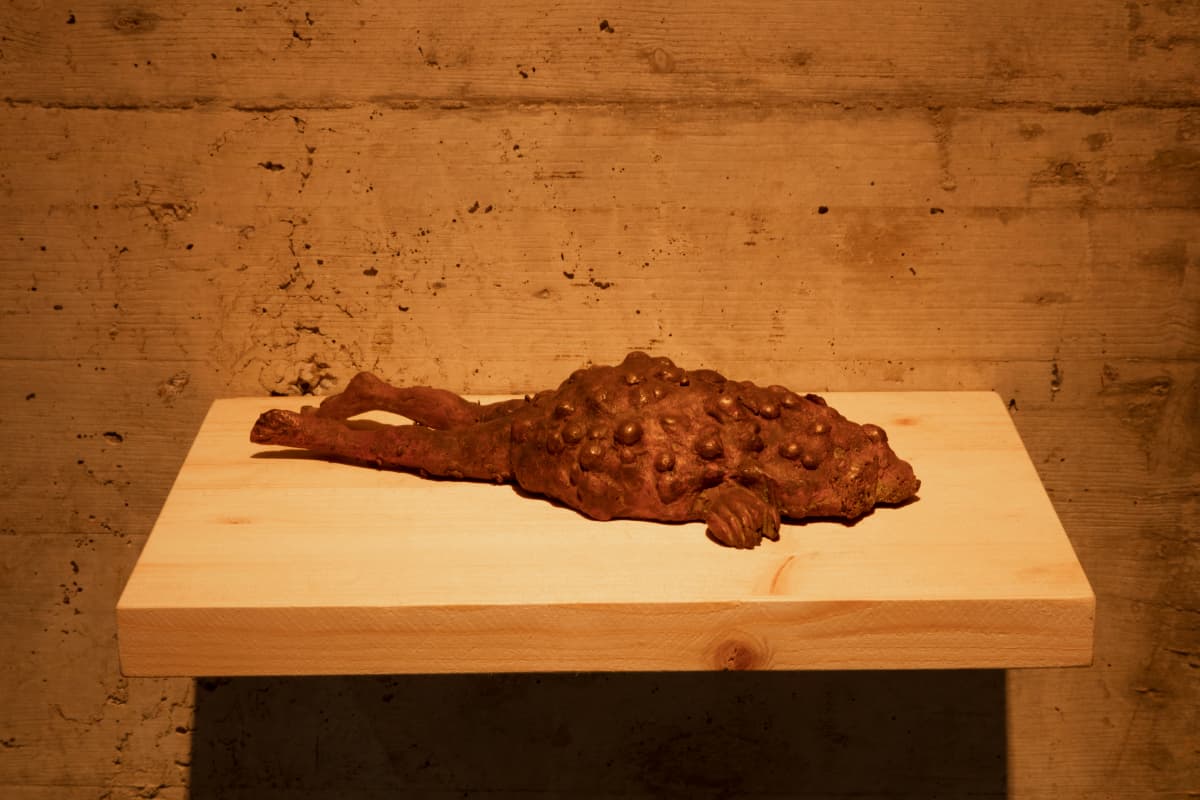 A textured, rust-colored sculpture of a reclining figure on a wooden shelf against a rough, beige wall.