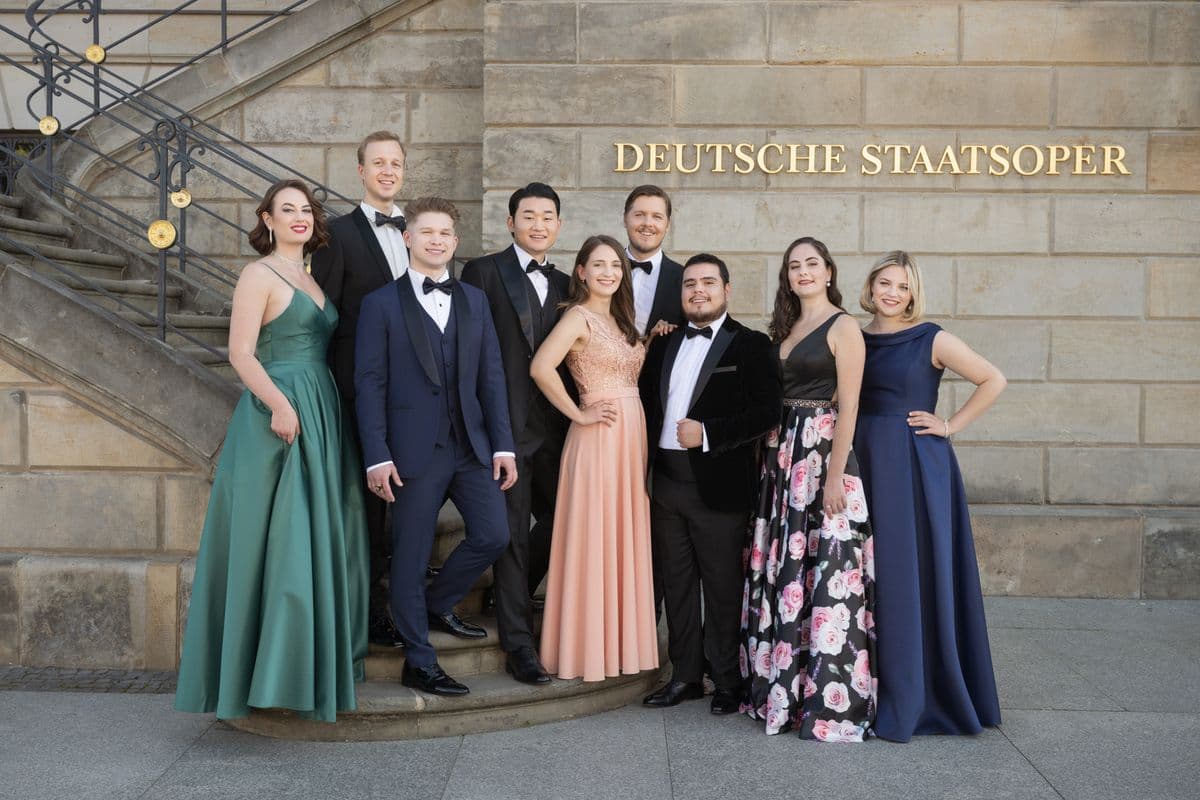 A group of nine people in formal attire pose in front of the Deutsche Staatsoper building.