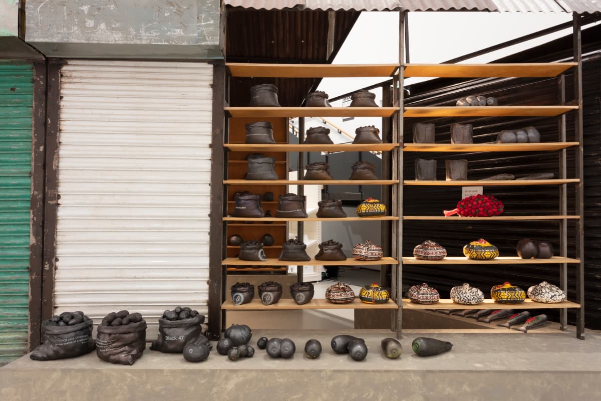 Shelves displaying various clay sculptures resembling shoes, fruits, and other objects in an outdoor market setting.