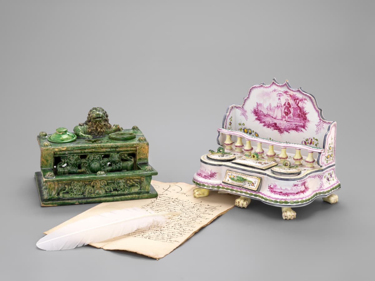 Zwei verzierte Tintenfässer: eines grün mit aufwendigen Schnitzereien, das andere weiß und rosa mit malerischen Motiven, daneben eine Feder und ein handgeschriebener Brief.