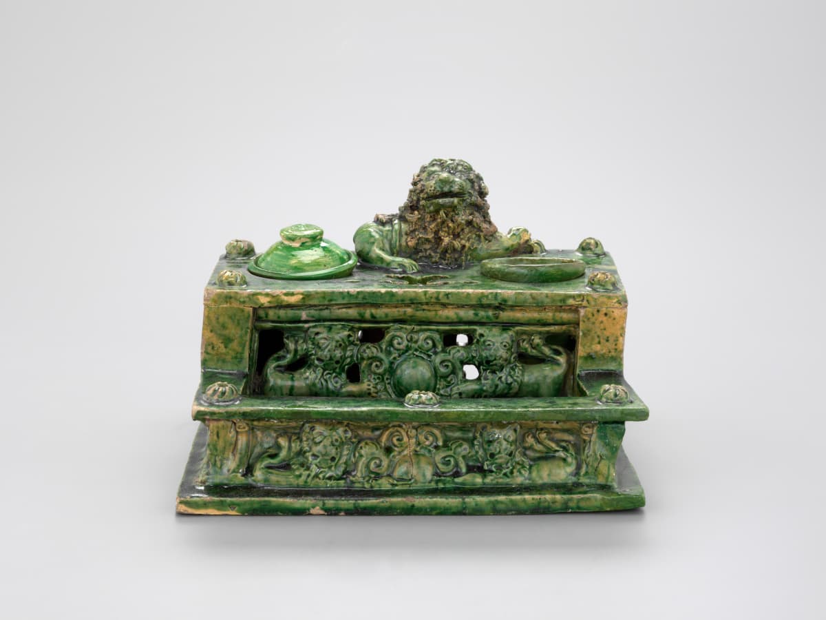 Grün glasiertes Keramiktintenfass mit komplizierten Mustern und einer Löwenfigur oben drauf, vor einem schlichten Hintergrund.