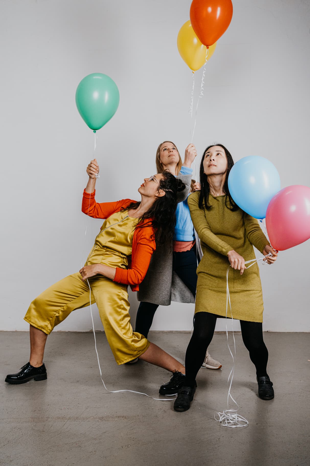 Drei Personen halten bunte Luftballons in kreativen Posen vor einem schlichten Hintergrund. Sie tragen farbenfrohe, legere Kleidung.