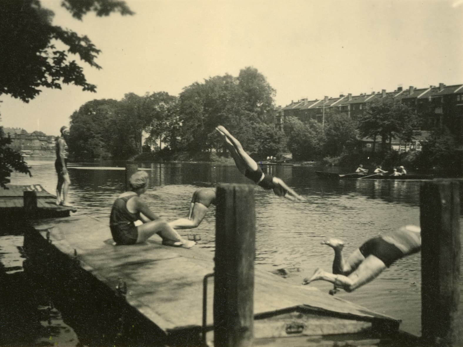 Black and white photograph of people on a bathing jetty by a river. Two people are jumping into the water, another is sitting on the jetty and one person is standing on the edge. Trees, houses and rowing boats with people on the river can be seen in the background. The scene looks lively and summery.