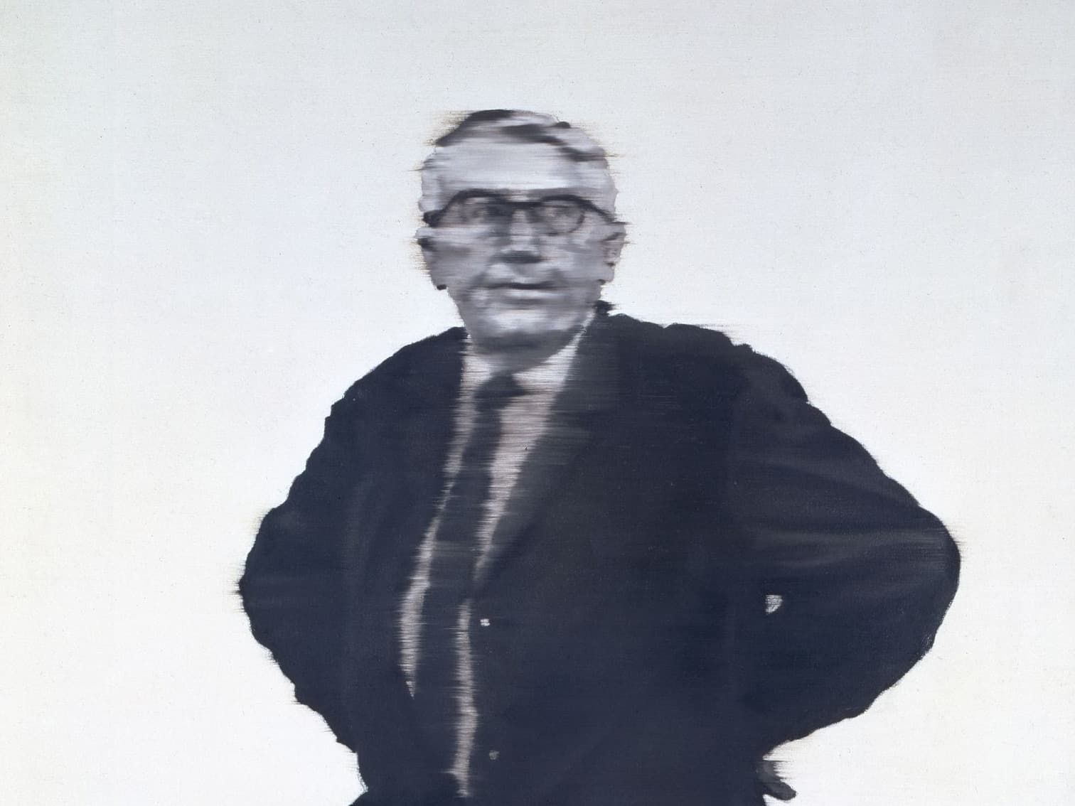 Unscharfes Bild eines Mannes im Anzug mit Brille, der mit den Händen in den Hüften vor einem schlichten Hintergrund steht.