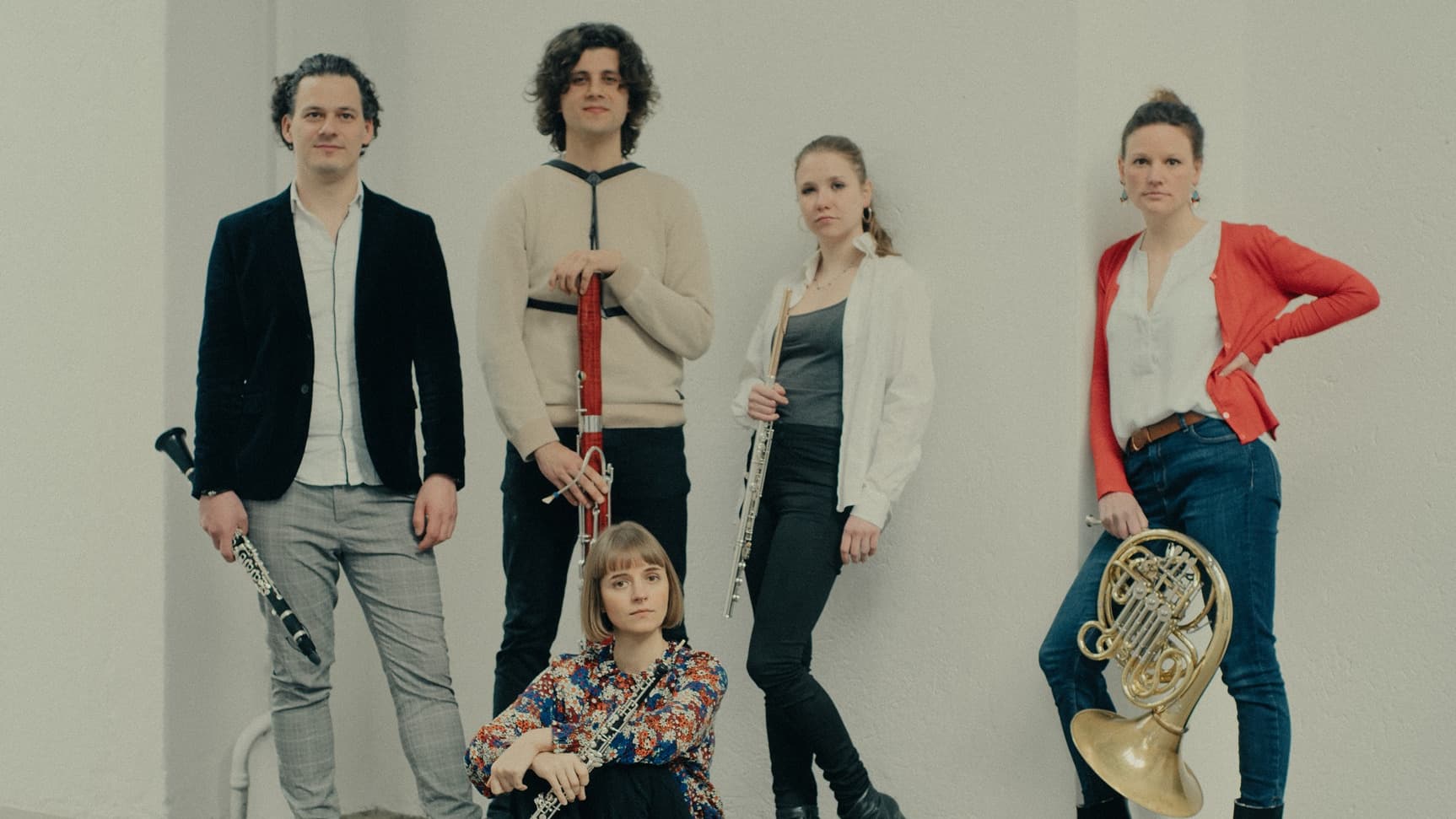 Five musicians pose with instruments, including clarinets, a bassoon, a flute, and a French horn, against a plain backdrop.