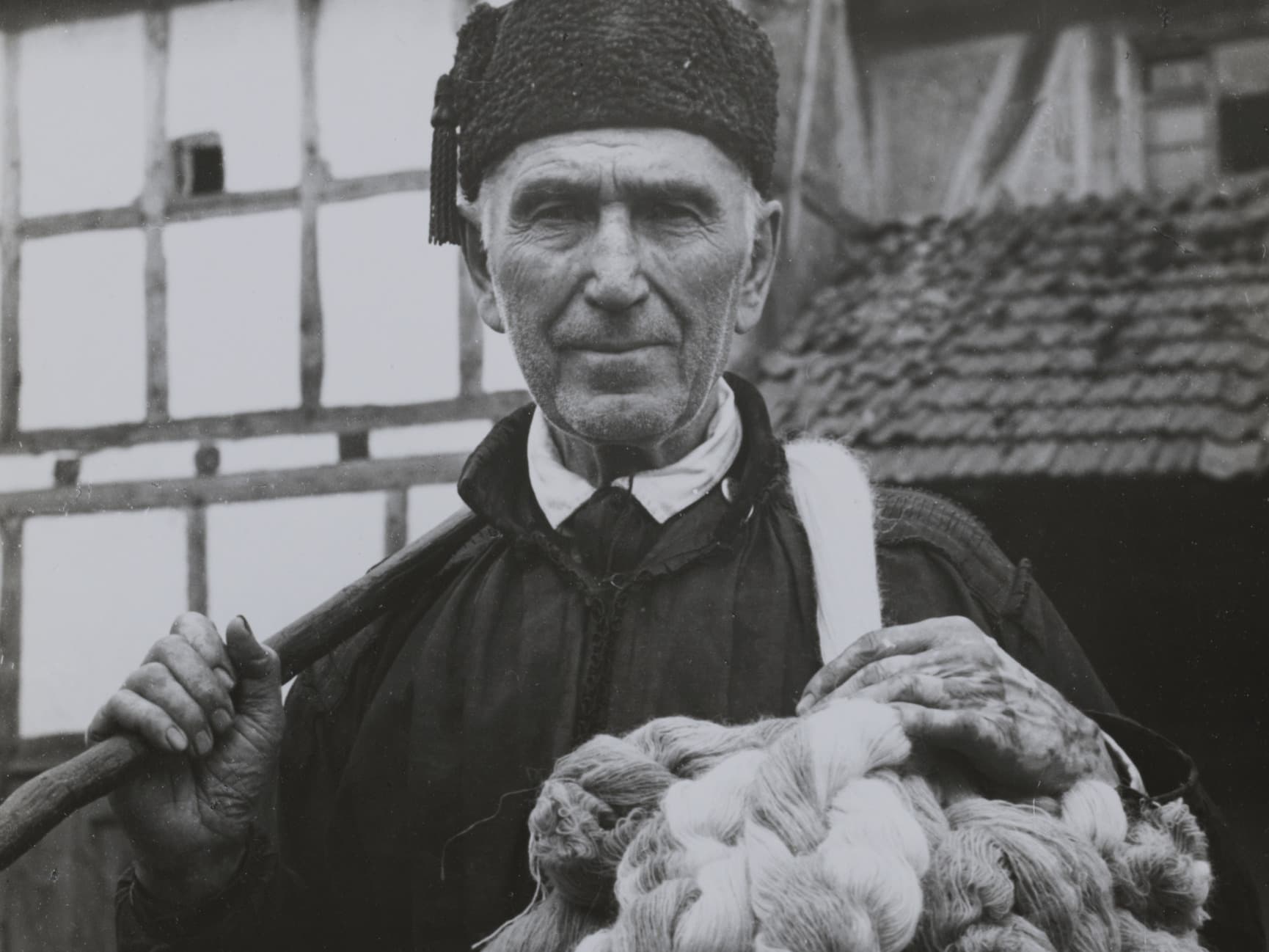 Ein älterer Mann in traditioneller Kleidung hält ein Bündel Garn und steht vor einem rustikalen Gebäude mit Fachwerk.