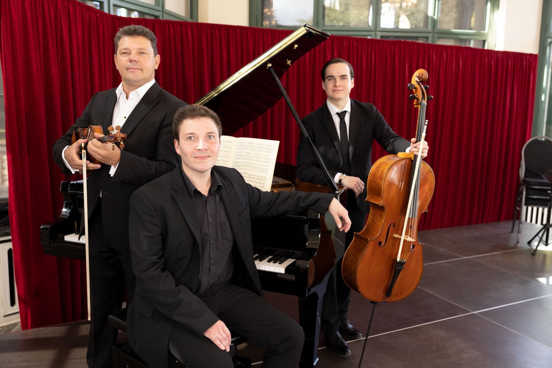 Three musicians in suits pose with a violin, piano, and cello in front of a red curtain.