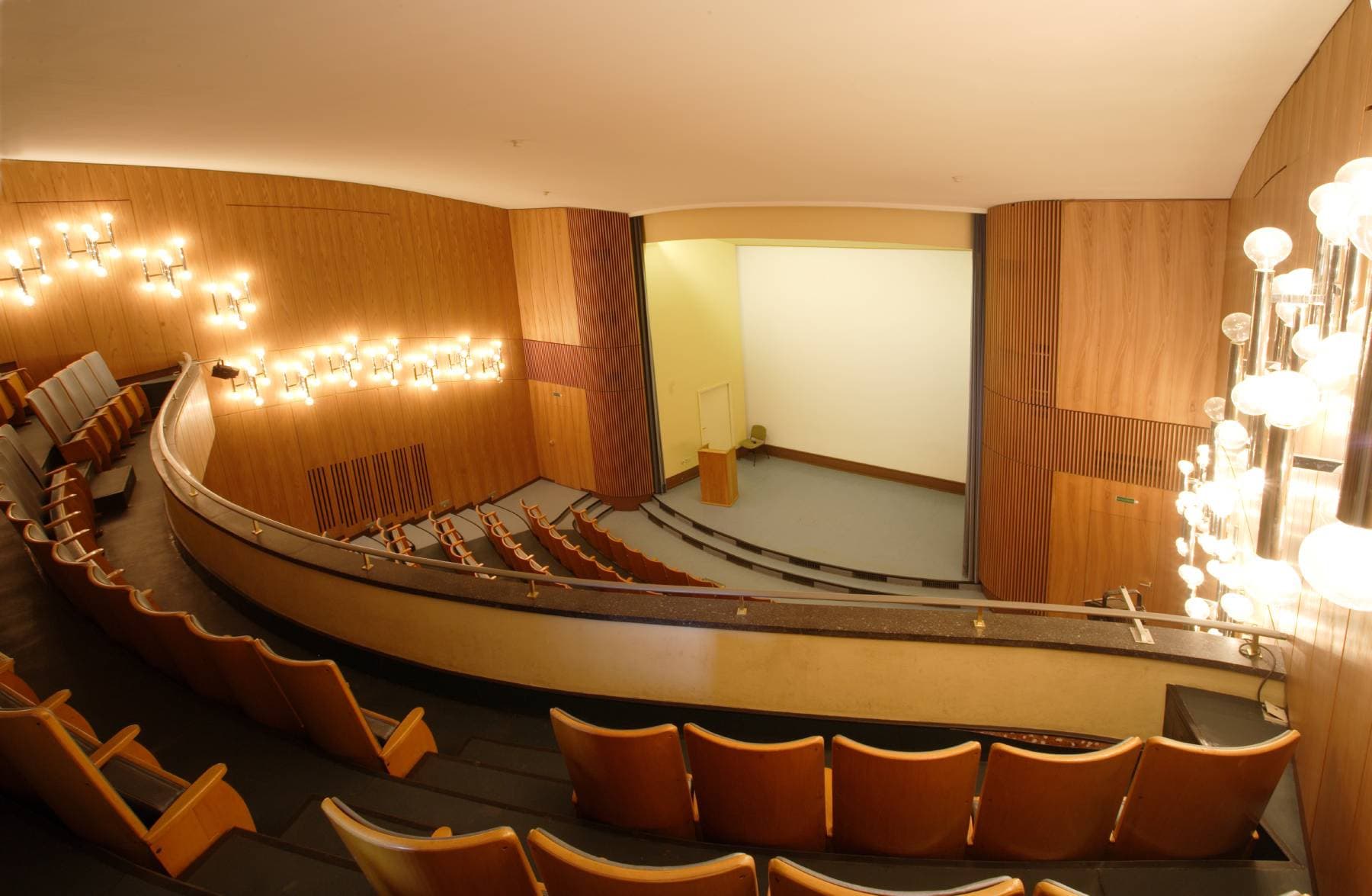 Leerer Zuschauerraum mit geschwungenen Sitzen, Holzwänden und warmer Beleuchtung, gegenüber einer kleinen Bühne mit Podium und großer Leinwand.