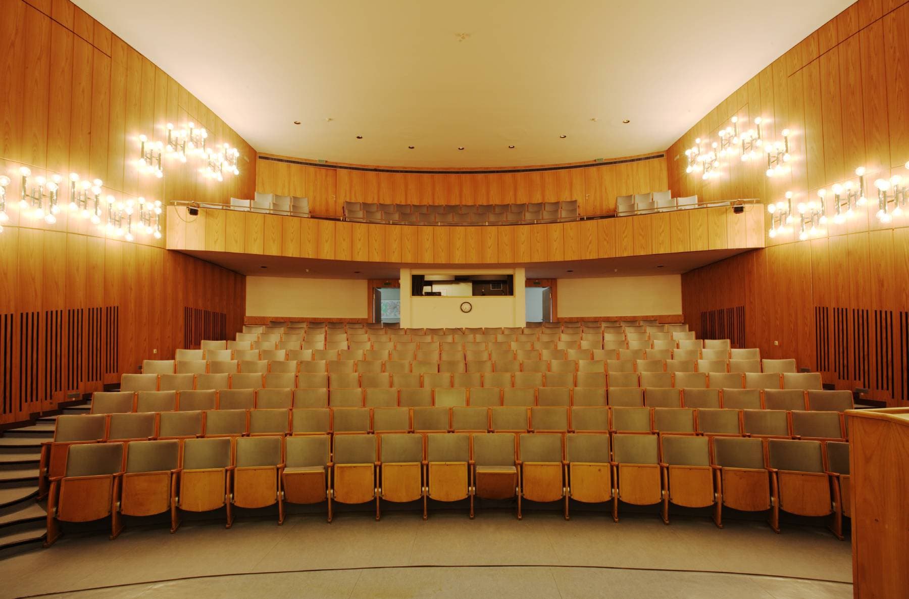 Leerer Zuschauerraum mit Holzwänden, Sitzreihen und warmer Beleuchtung. Balkonbereich und eine kleine Uhr an der Rückwand.