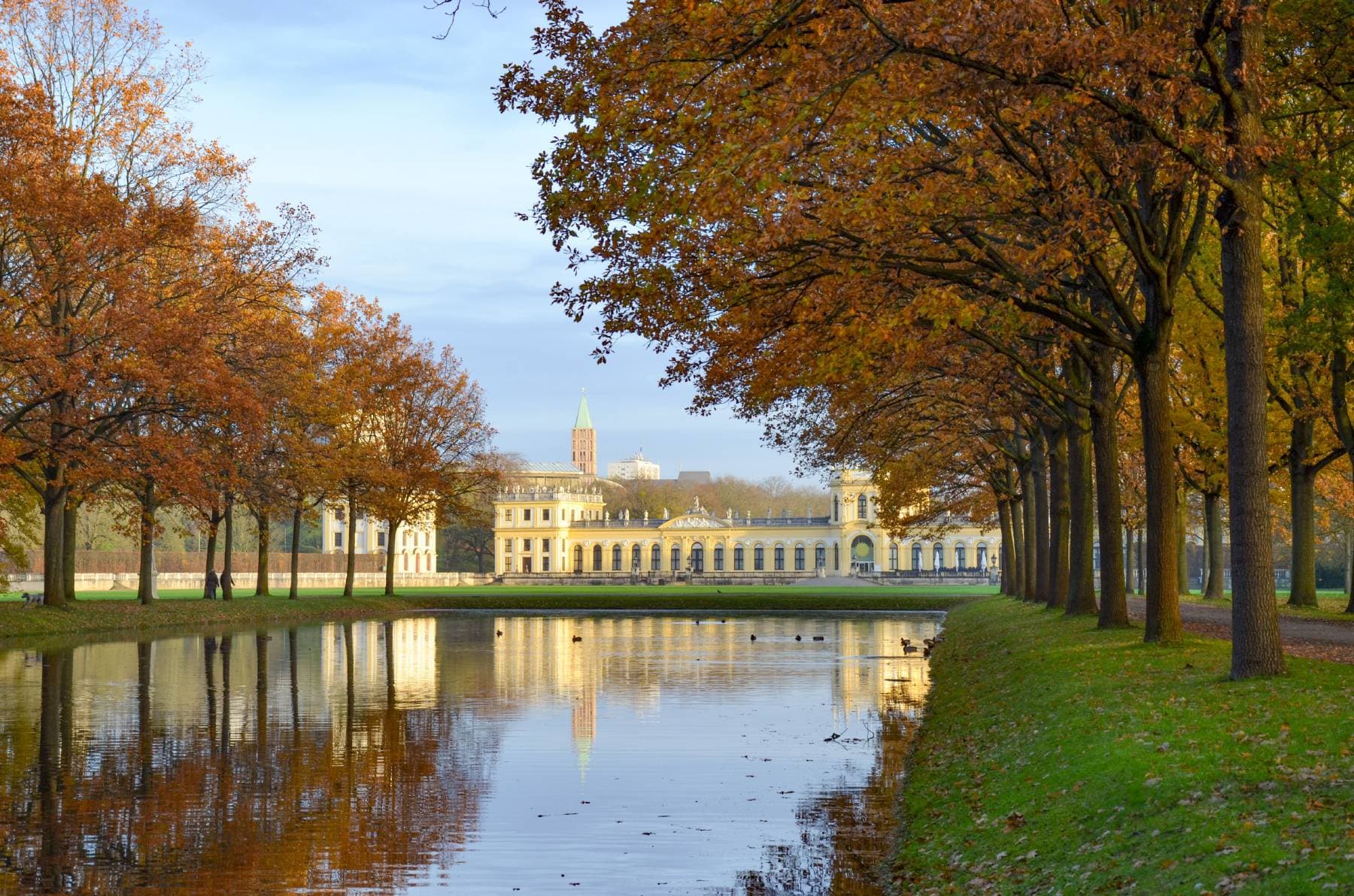 Ein von Bäumen gesäumter Weg, in dem sich das Herbstlaub in einem ruhigen Fluss spiegelt, führt zu einem weit entfernten, prachtvollen Gebäude unter blauem Himmel.
