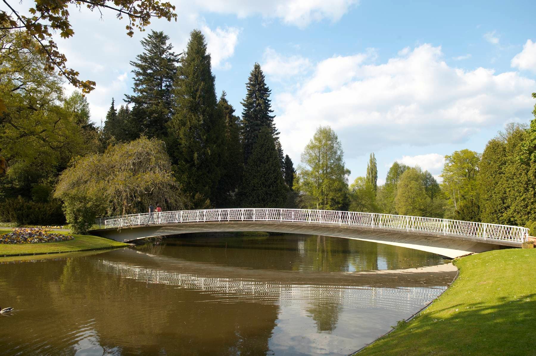 Eine weiße Bogenbrücke überspannt einen ruhigen Fluss in einem Park, umgeben von üppigem Grün und hohen Bäumen unter einem teilweise bewölkten Himmel.