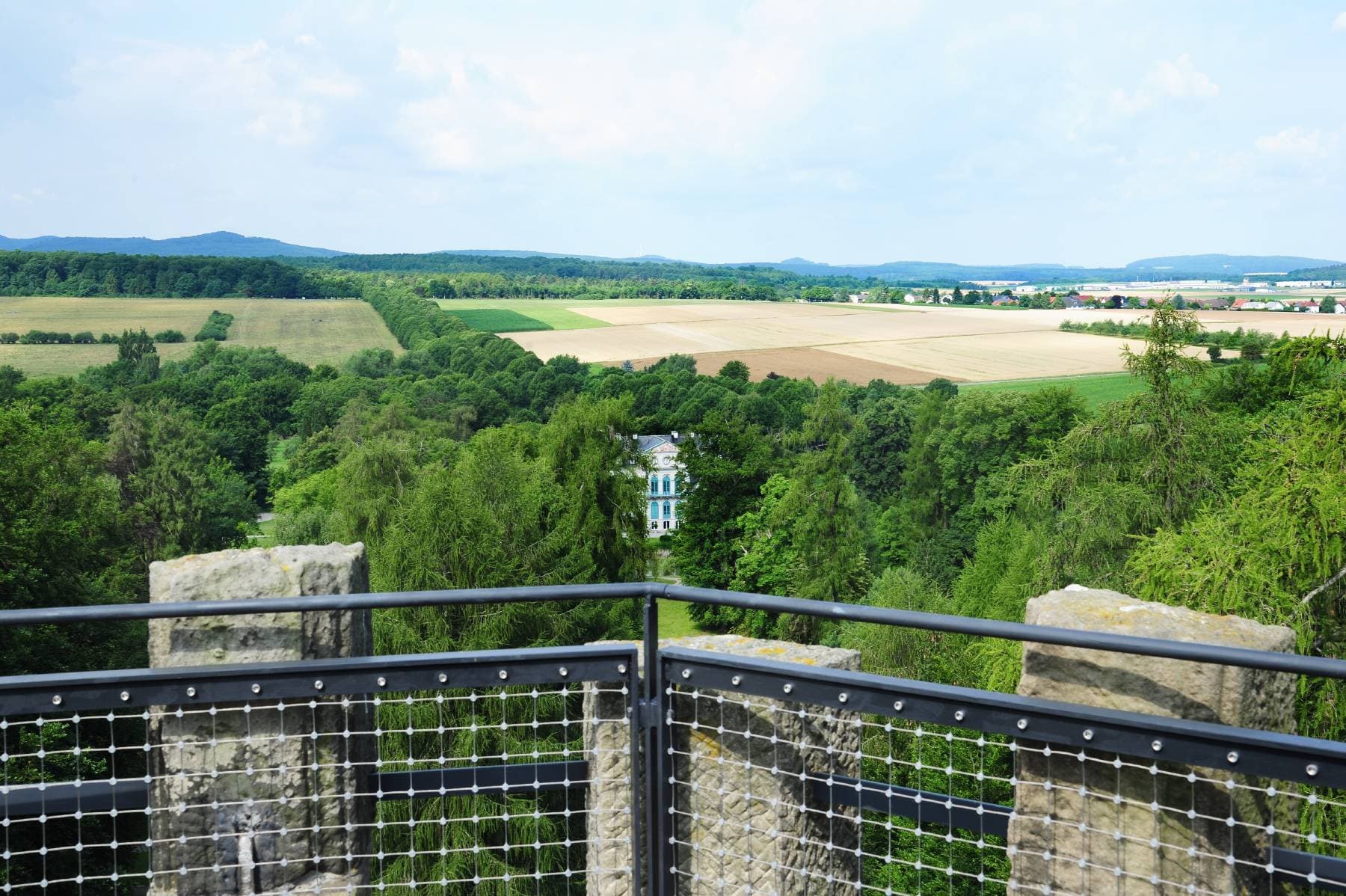 Blick von einer Steinterrasse auf eine üppig grüne Landschaft mit Feldern, Bäumen und einem entfernten Gebäude, das teilweise durch Laub verdeckt ist.