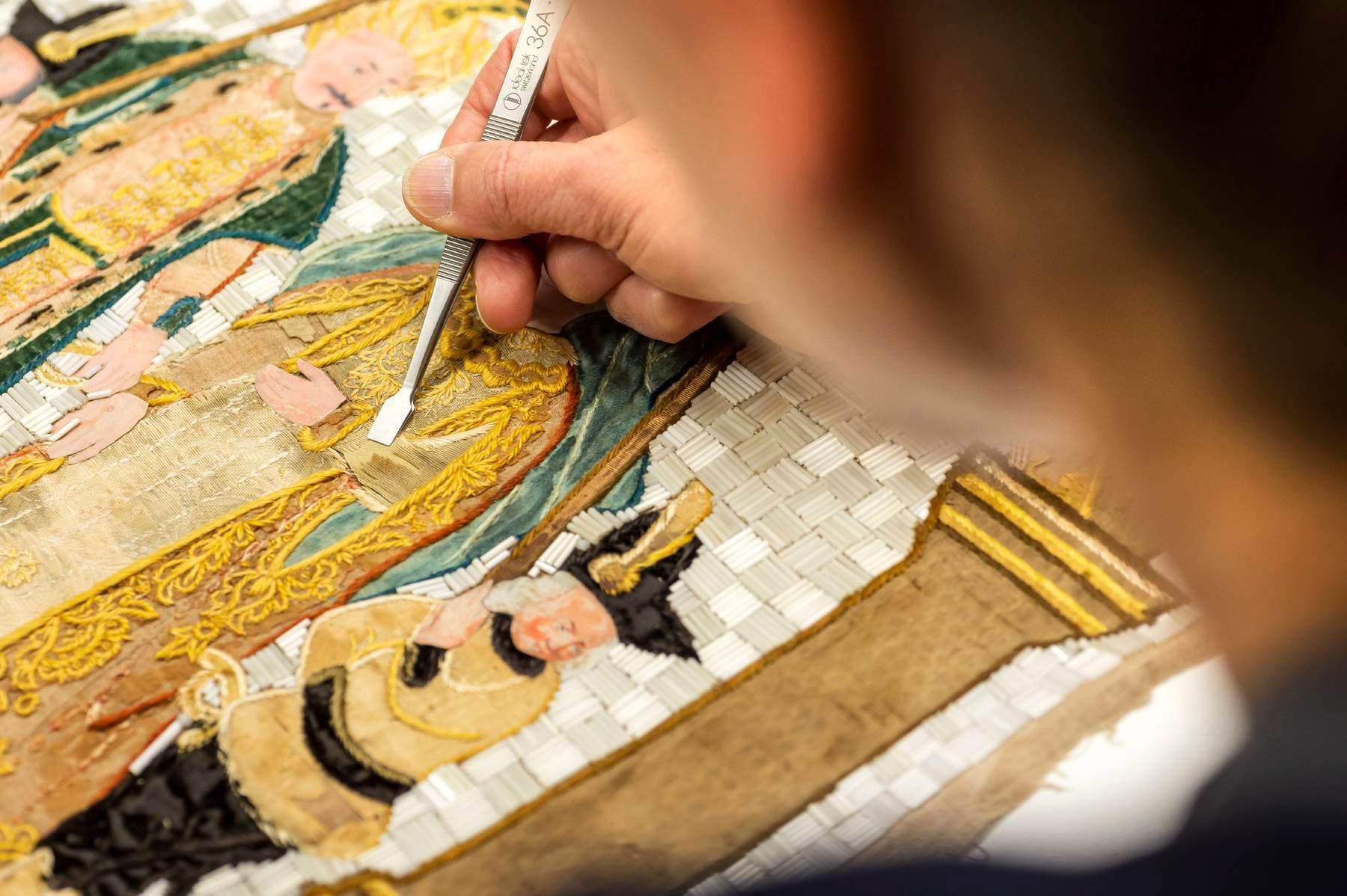 Eine Person restauriert sorgfältig mit einem kleinen Werkzeug einen detailreichen, farbenfrohen Wandteppich und konzentriert sich dabei auf komplizierte Goldmuster und historische Figuren.