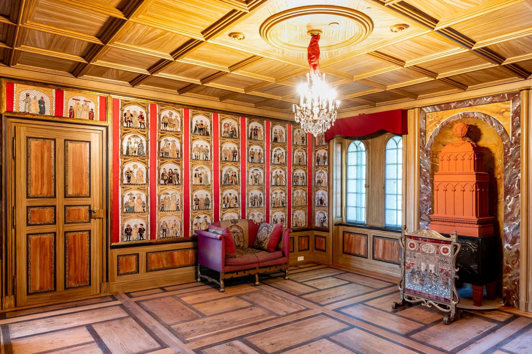 Reich verzierter Raum mit holzgetäfelter Decke, Kronleuchter, dekorativen Wänden, gewölbtem Kamin, rotem Sofa und großen Fenstern mit roten Vorhängen.