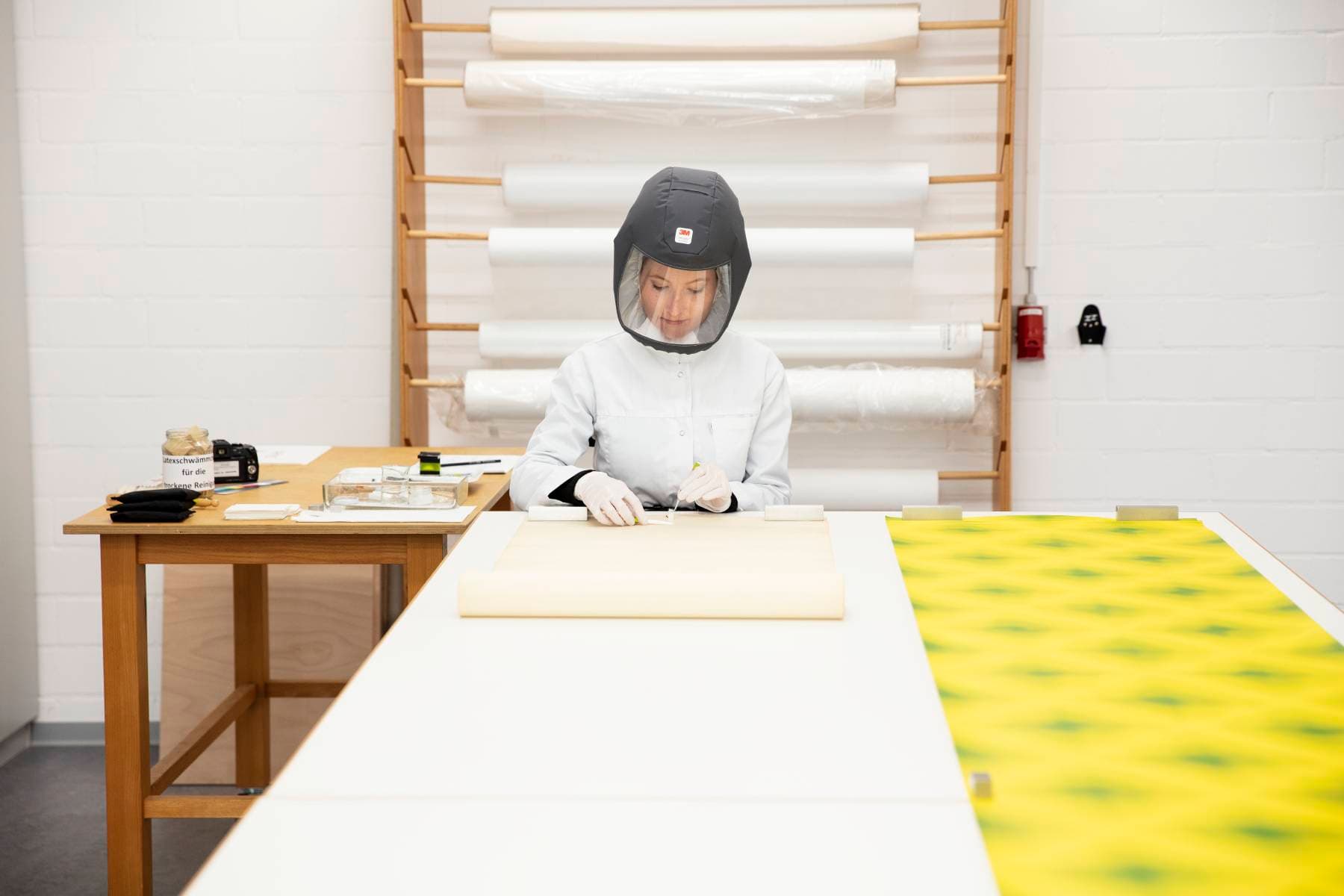 Eine Person in Schutzkleidung arbeitet an einem Tisch mit Kunstmaterialien, umgeben von Papierrollen in einem hellen, ordentlichen Studio.