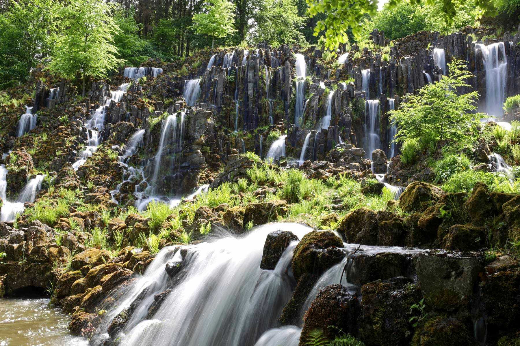 Breit gefächerter Wasserfall, der über viele Basaltsäulen hinabfällt, eingebettet in den grünen Bergpark