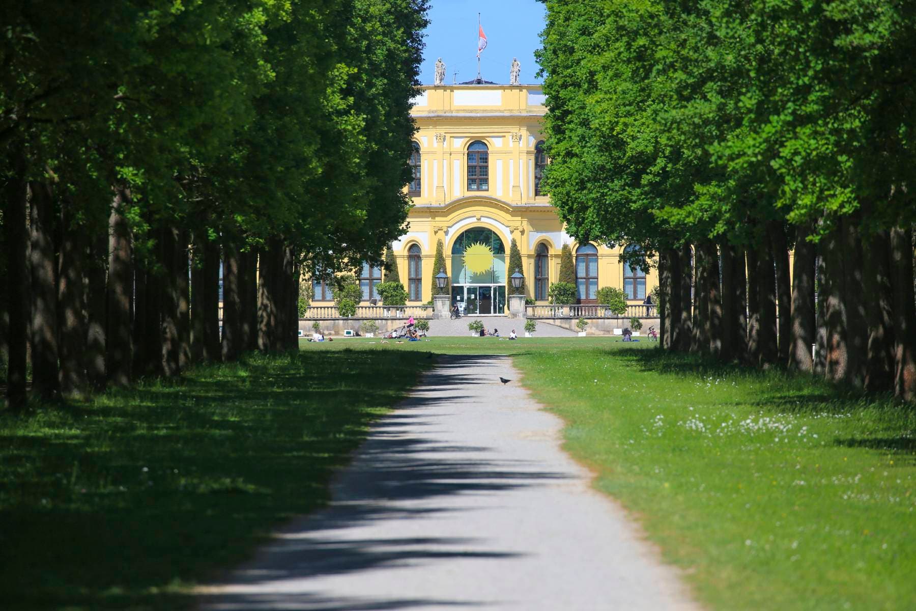 Von Bäumen gesäumter Weg, der zu einem großen gelben Gebäude mit gewölbtem Eingang führt, umgeben von üppigem Grün unter einem klaren blauen Himmel.