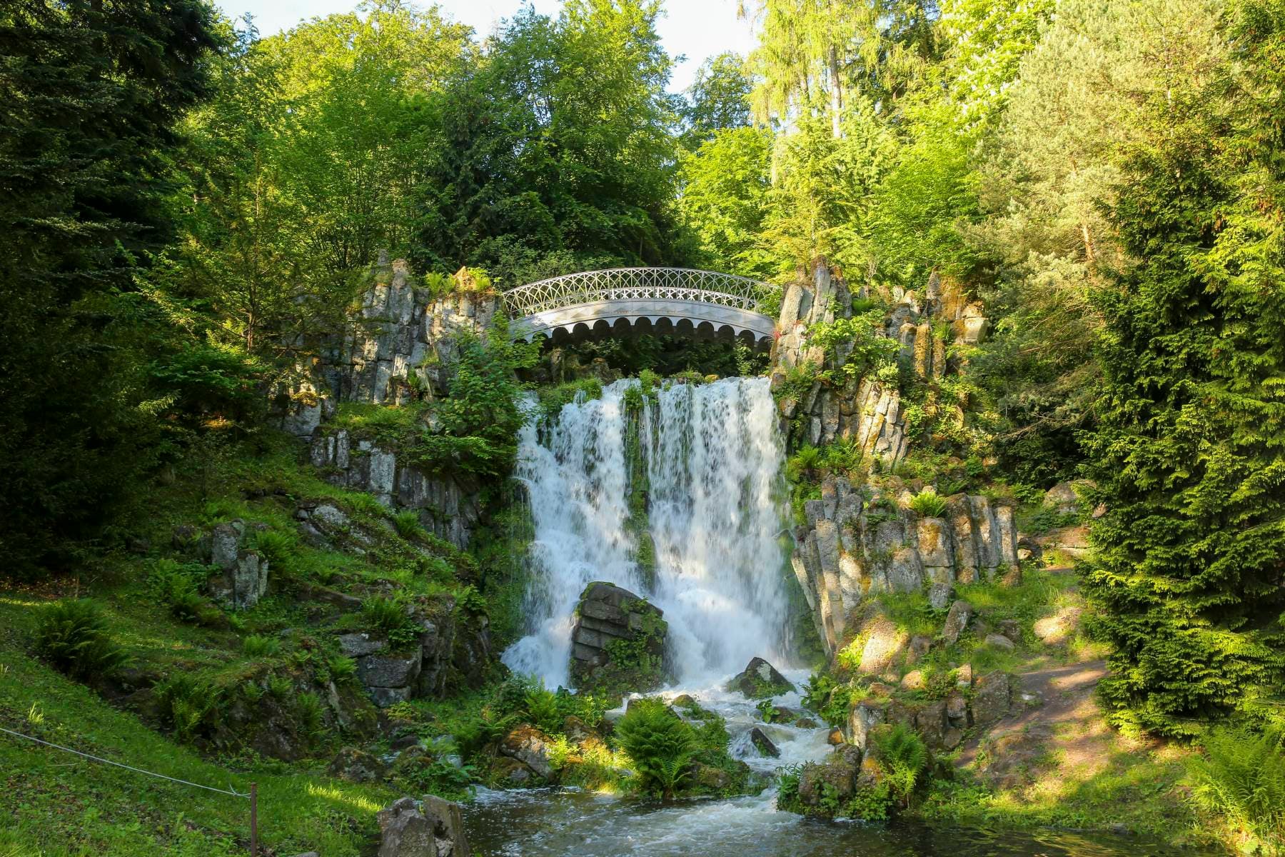 Wasserfall mit darüber verlaufender Brücke, eingebettet in den grünen Bergpark