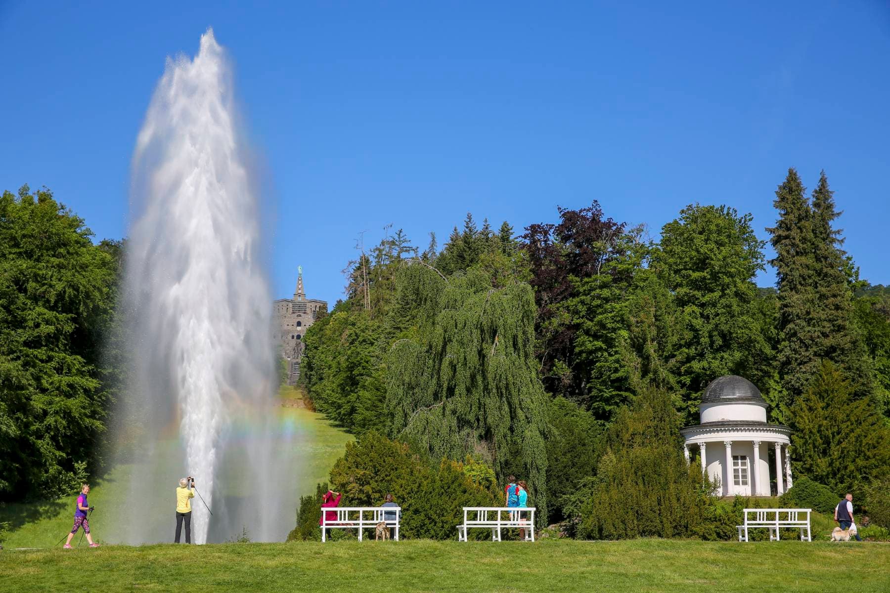 Ein großer Brunnen spritzt Wasser hoch und erzeugt einen Regenbogen, mit Menschen in der Nähe und einem kleinen Pavillon, umgeben von Bäumen unter einem klaren blauen Himmel.