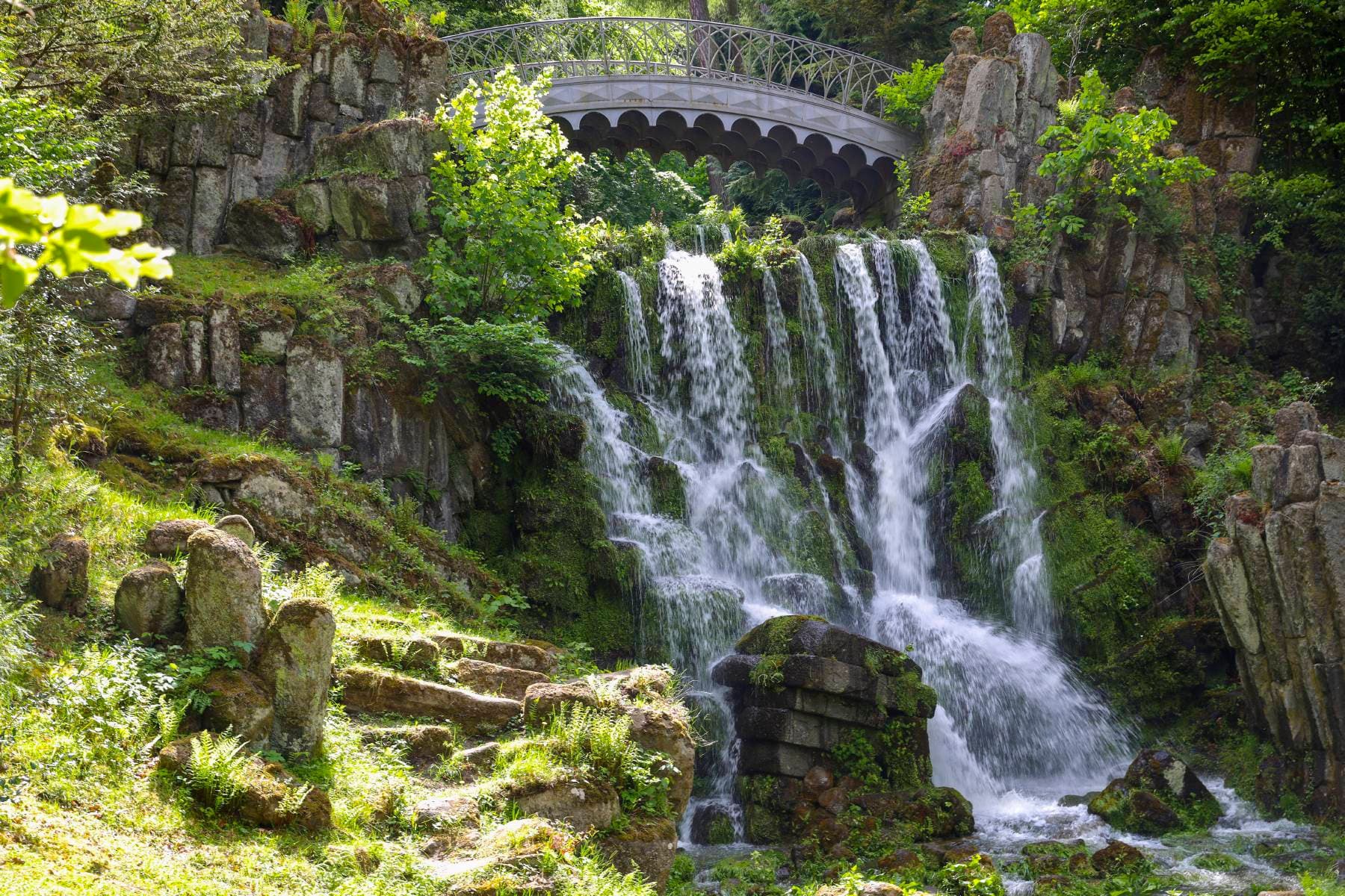 Wasserfall mit darüber verlaufender Brücke, eingebettet in den grünen Bergpark