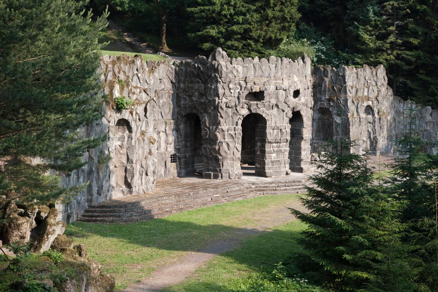 Steinstruktur mit gewölbten Eingängen, umgeben von üppigen grünen Bäumen und Gras, die einer antiken Ruine oder Grotte ähnelt.