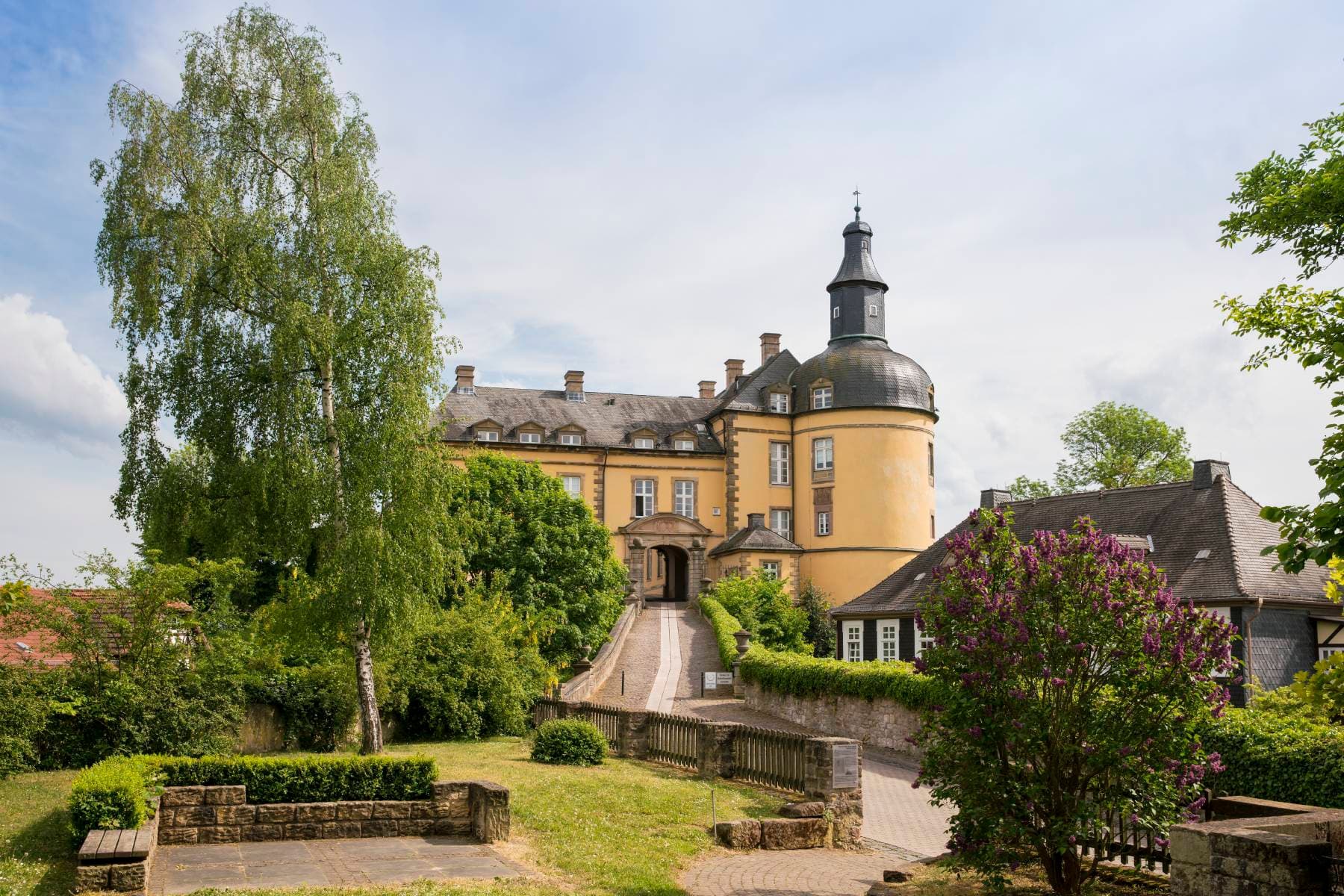 Historisches gelbes Schloss mit Turm, umgeben von üppigem Grün und einem Kopfsteinpflasterweg, unter einem teilweise bewölkten Himmel.