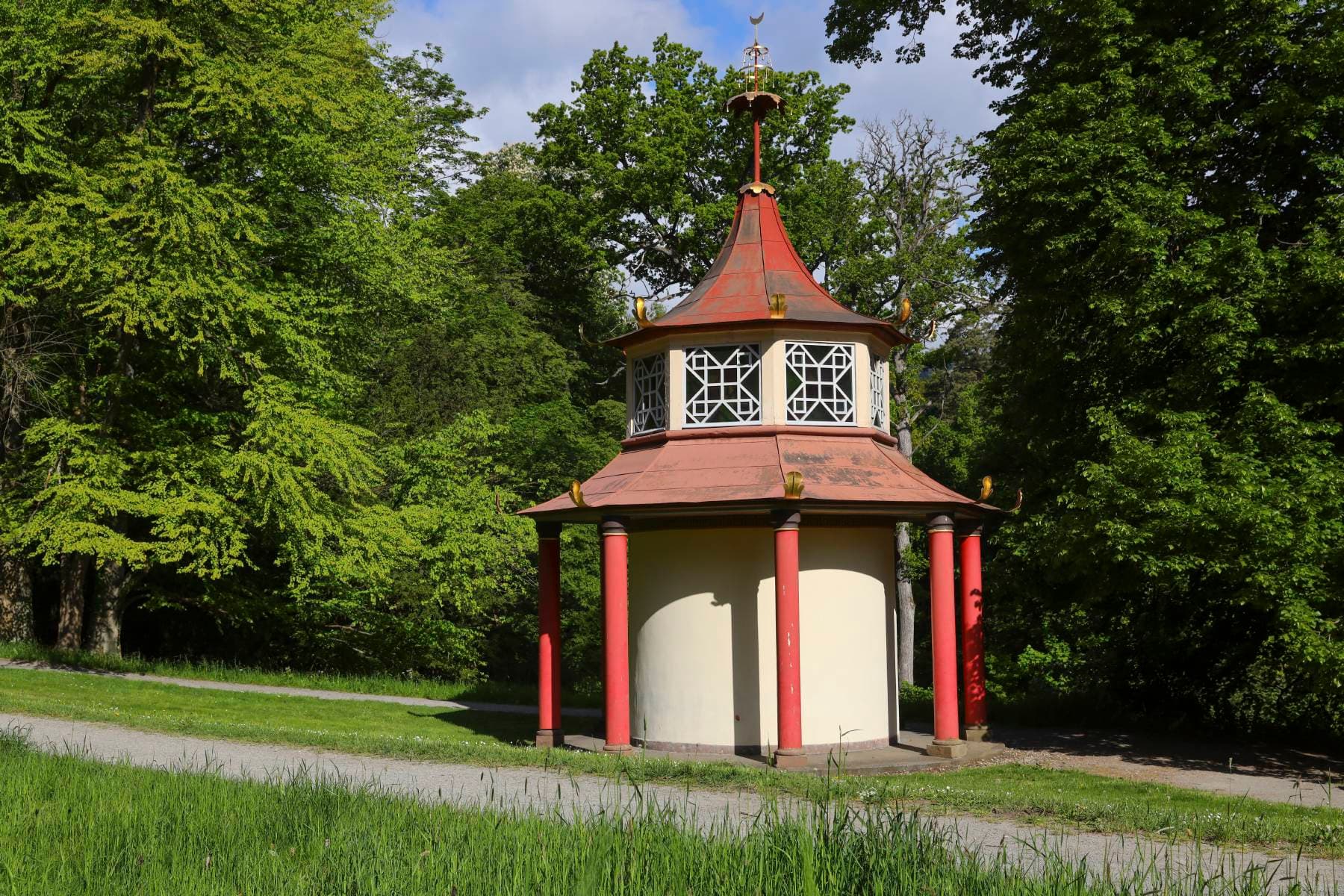 Ein kleiner, achteckiger Pavillon mit rotem Dach und Säulen, umgeben von üppigen grünen Bäumen und Gras an einem sonnigen Tag.