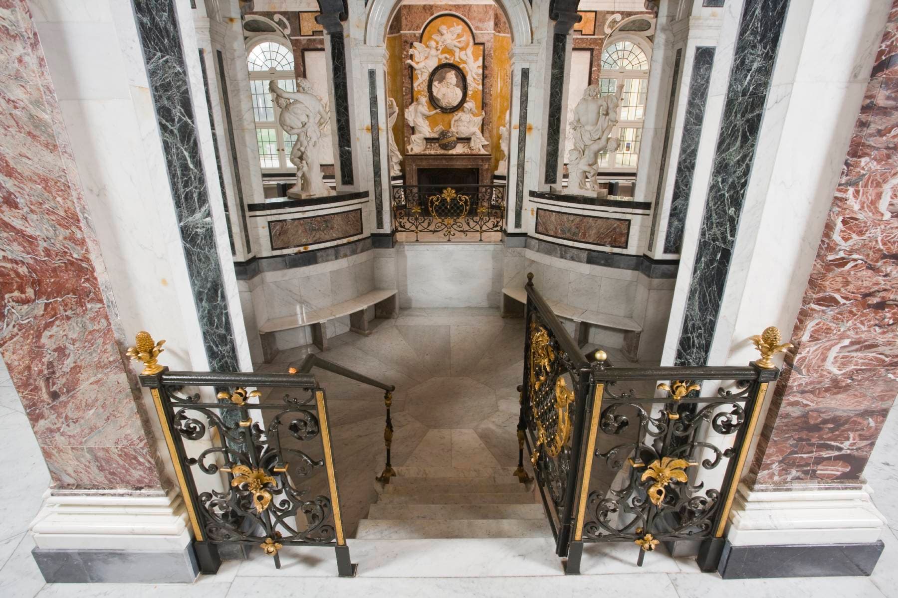 Eine kunstvolle Marmortreppe mit dekorativen schmiedeeisernen Geländern und Statuen führt zu einer unteren Ebene in einem großen, klassischen Gebäude.