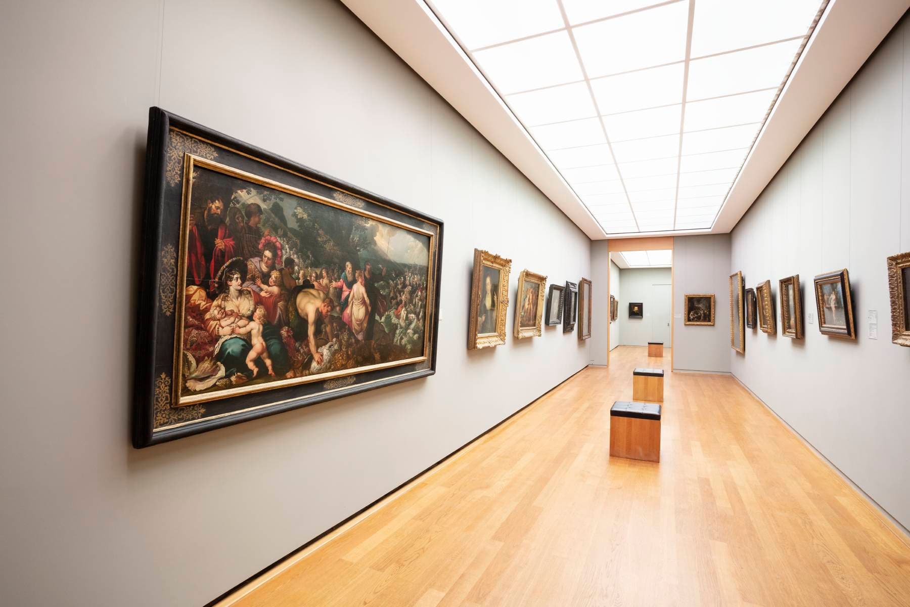 Innenraum einer Kunstgalerie mit Gemälden an hellgrauen Wänden, Holzboden und einer Bank. Helle Deckenbeleuchtung erhellt den Raum.