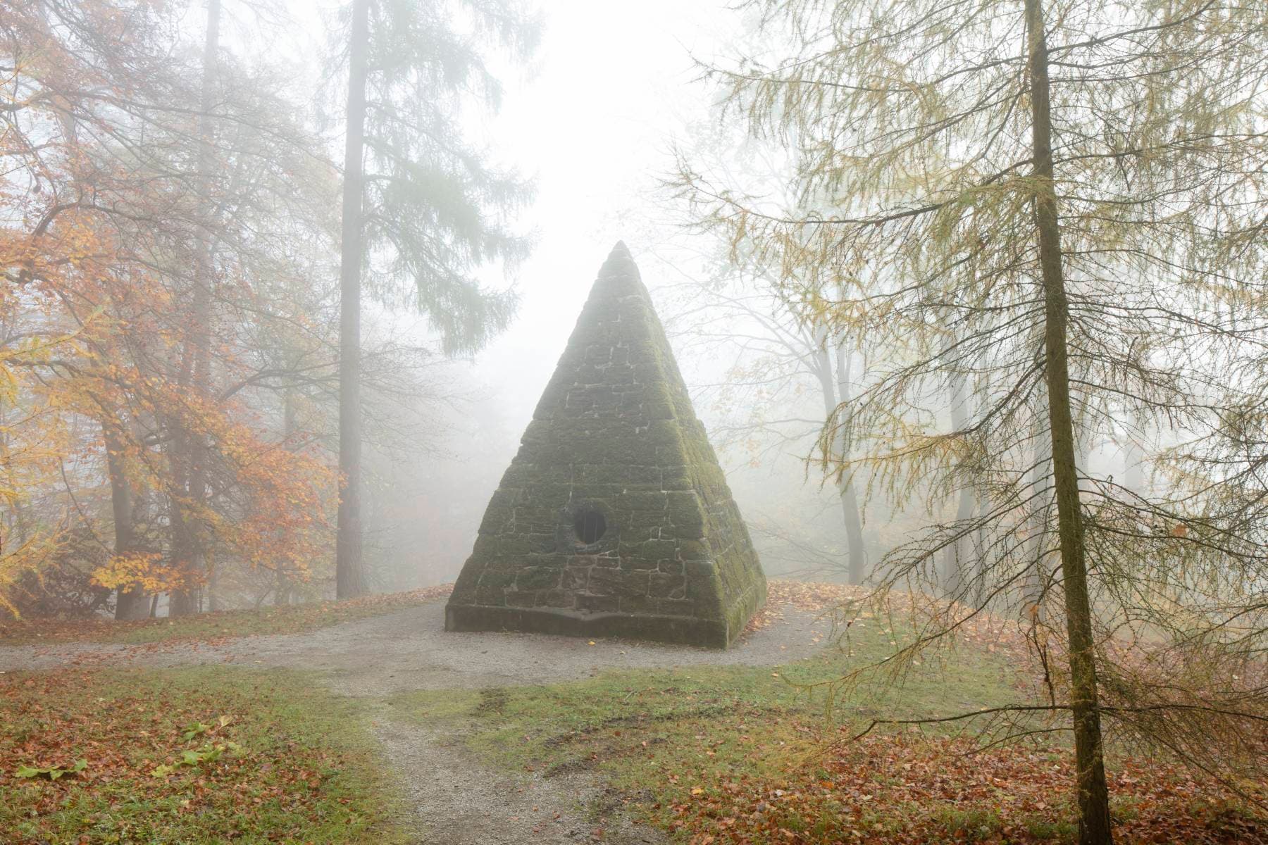 Eine moosbedeckte Steinpyramide steht in einem nebligen Wald, umgeben von Herbstbäumen mit orangefarbenen und gelben Blättern.