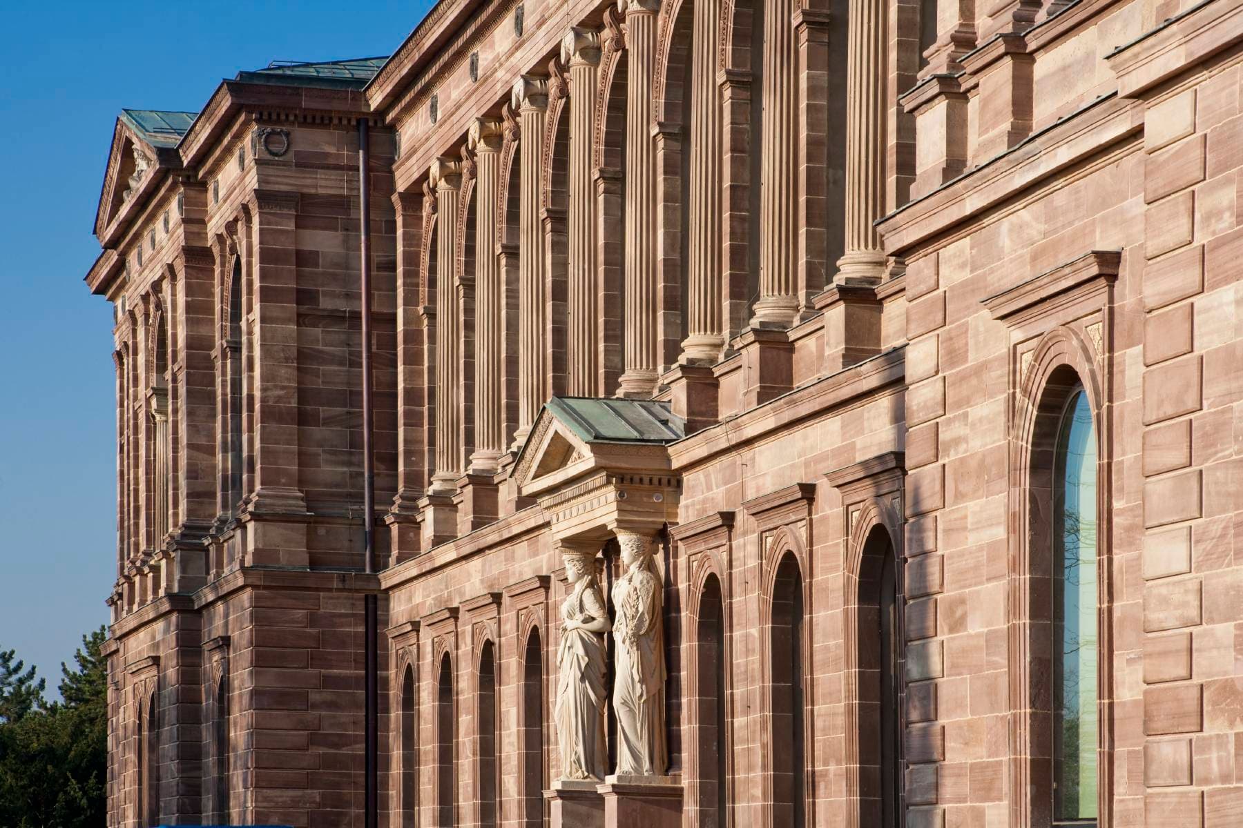 Seitenansicht eines historischen Gebäudes mit hohen Bogenfenstern, verzierten Säulen und klassischen Statuen unter einem klaren blauen Himmel.