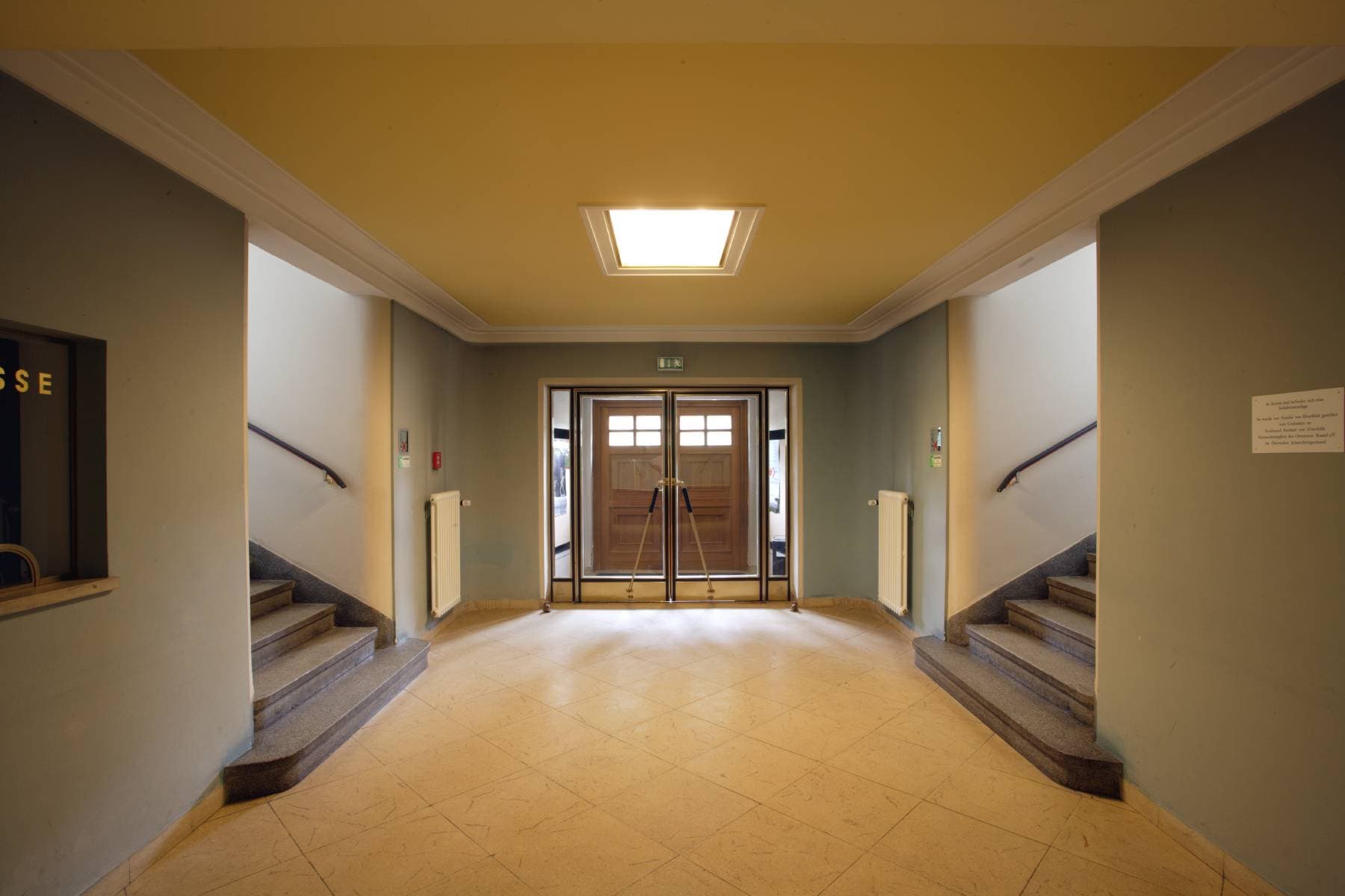 Geräumiges Foyer mit symmetrischen Treppen auf beiden Seiten, die zu Glas-Doppeltüren unter einer Deckenleuchte führen.