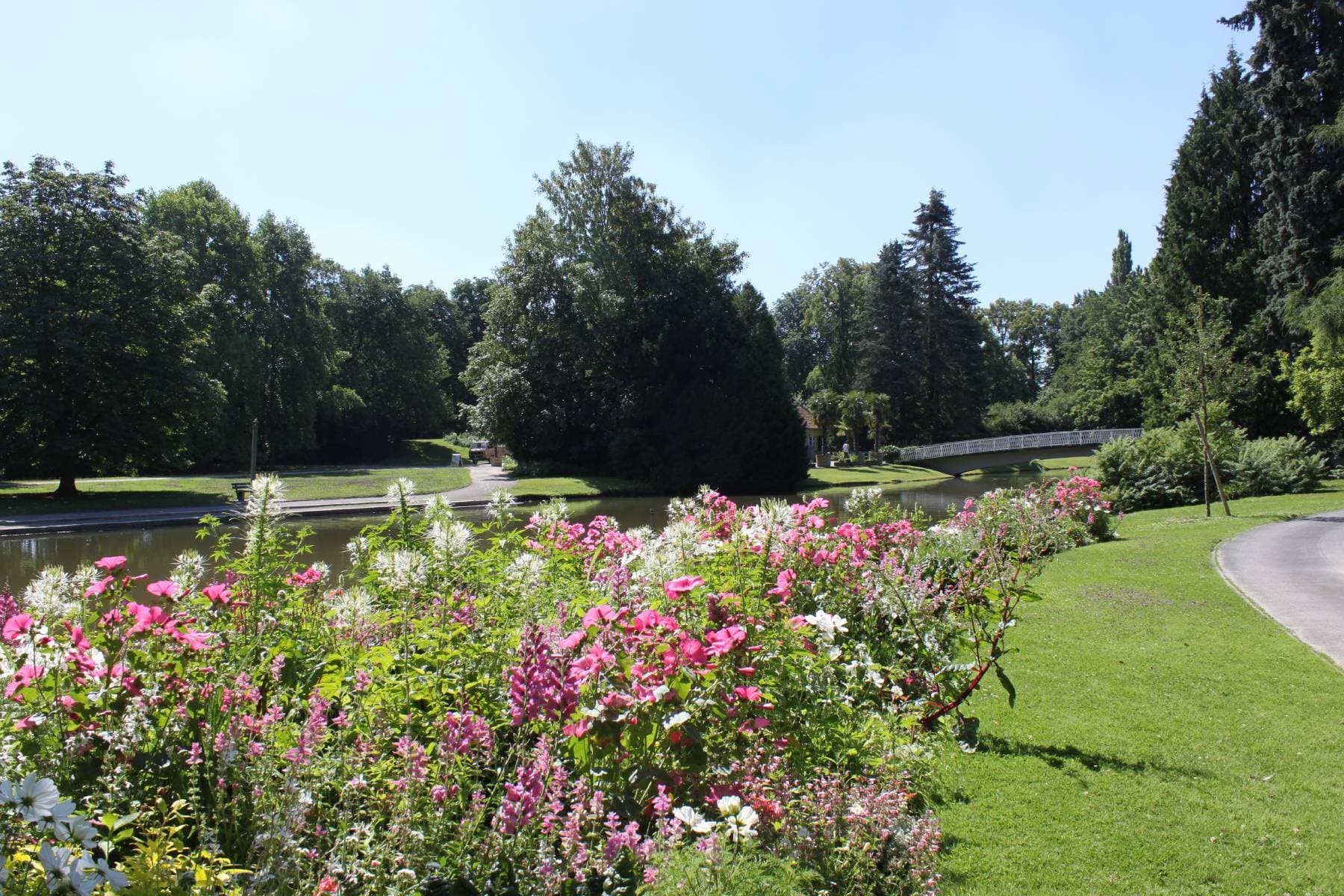 Ein malerischer Park mit leuchtenden Blumen im Vordergrund, einem gewundenen Pfad, Bäumen und einer kleinen Brücke über einem Teich unter einem klaren blauen Himmel.