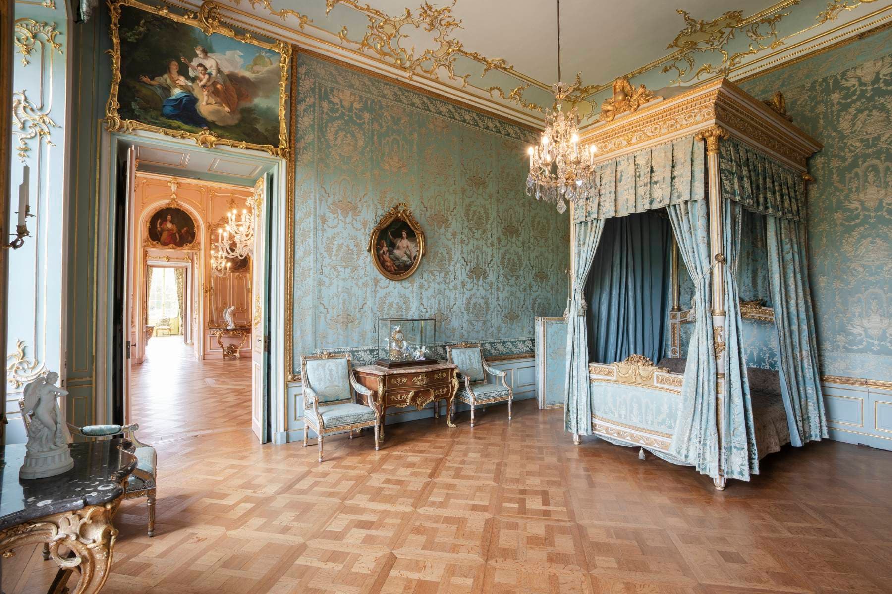 Elegantes Zimmer im Rokoko-Stil mit verzierten blauen Wänden, einem Himmelbett, Kronleuchtern und Parkettboden. Dekorative Gemälde und Möbel schmücken den Raum.