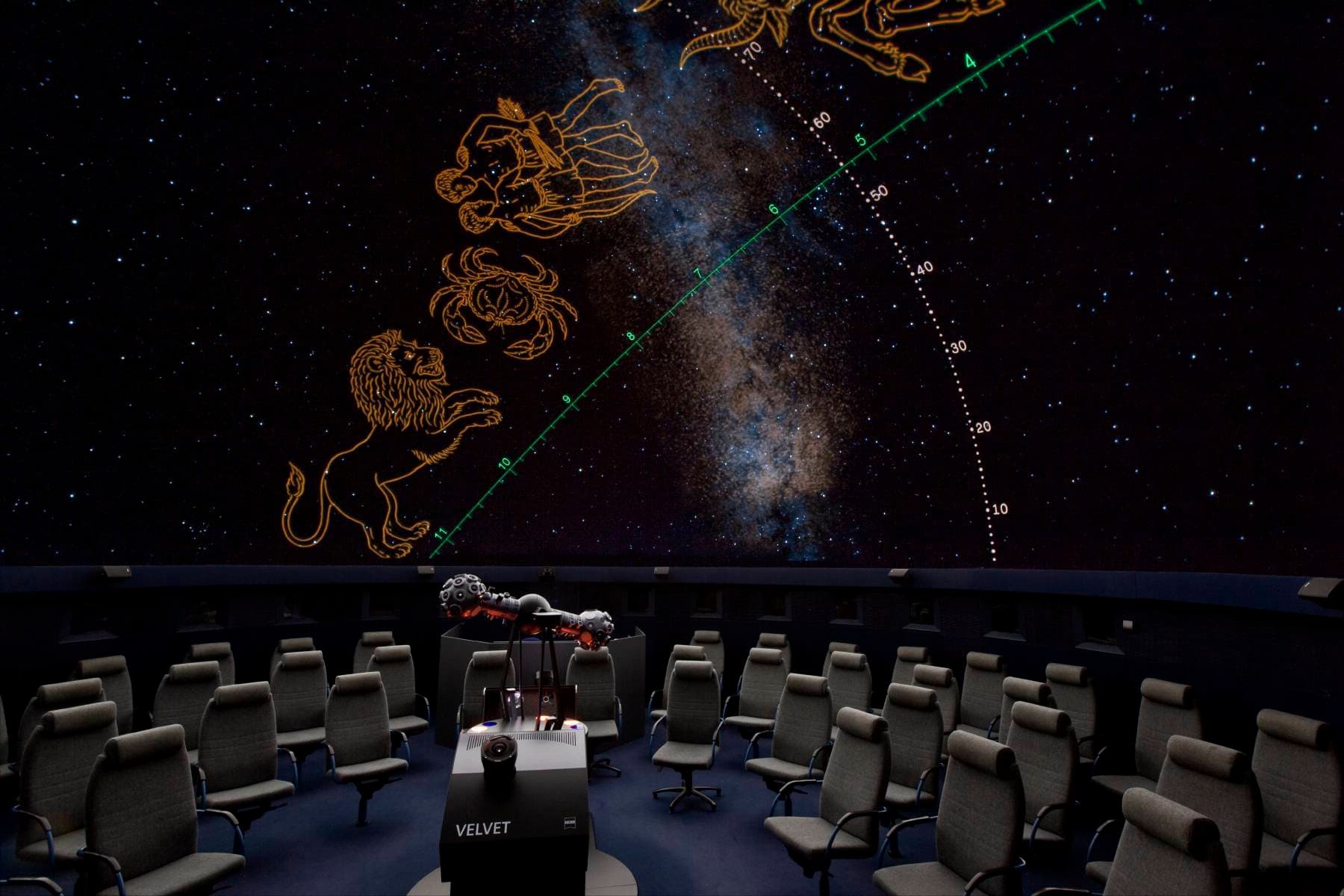 Planetariumsinnenraum mit Sitzreihen gegenüber einer Sternenkuppel, in der Sternbilder, darunter ein Löwe, vor dem Hintergrund der Milchstraße zu sehen sind.