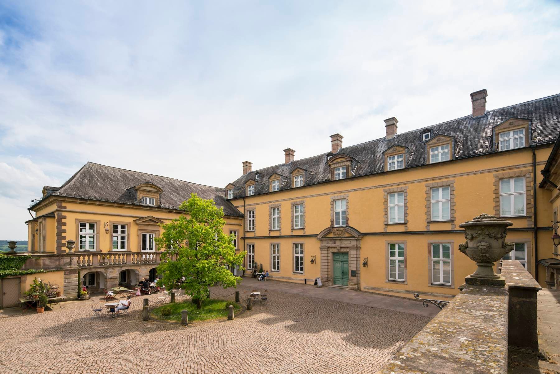 Ein großes, historisches gelbes Gebäude mit mehreren Fenstern, einem Innenhof mit Kopfsteinpflaster und einem grünen Baum in der Mitte unter einem teilweise bewölkten Himmel.