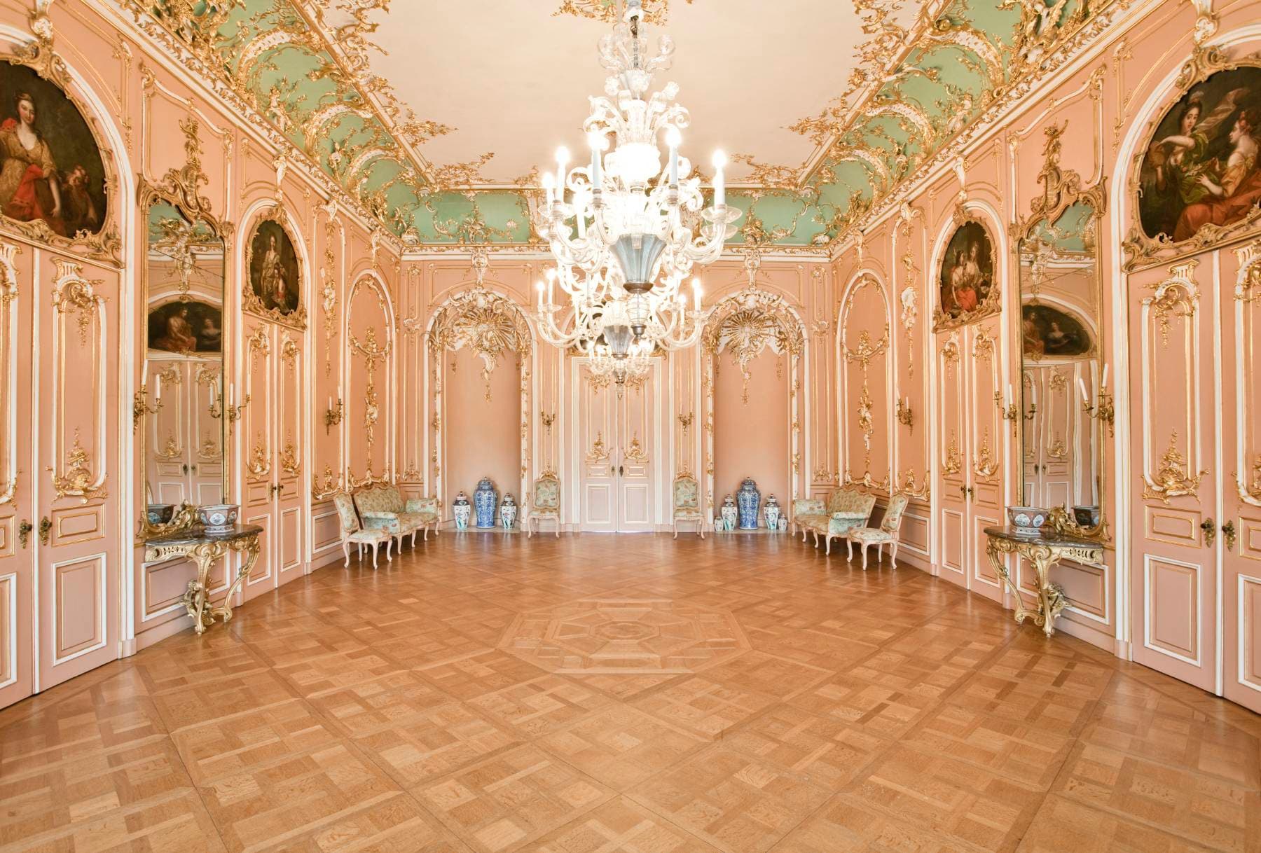 Reich verzierter Ballsaal mit rosa und goldenem Dekor, großem Kronleuchter, Parkettboden und Gemälden in gewölbten Rahmen an den Wänden.