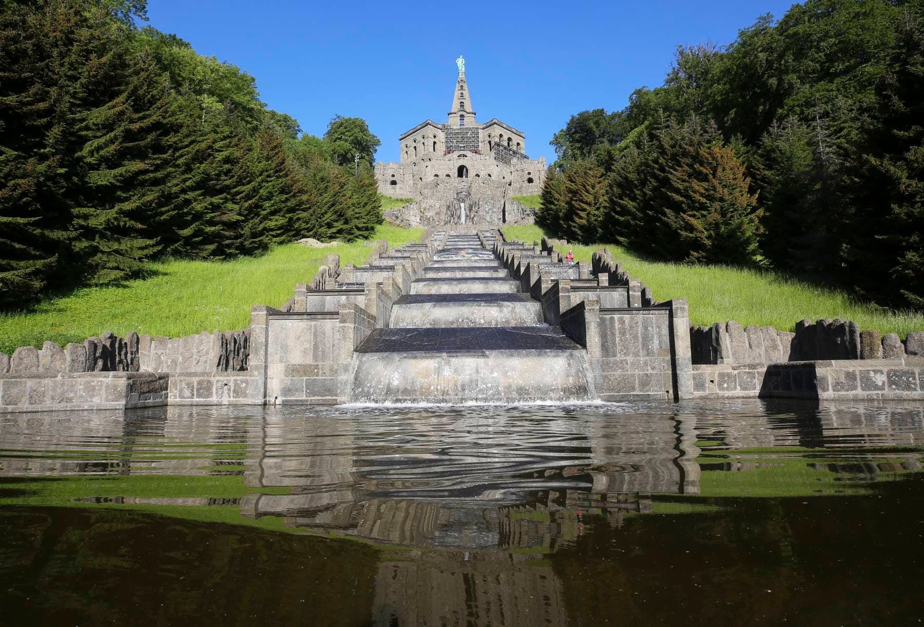 Blick über eine Wasserfläche und den großen Kaskaden bis hin zu einem Bauwerk, das von einer Pyramide und einer Herkulesstatue bekrönt wird.