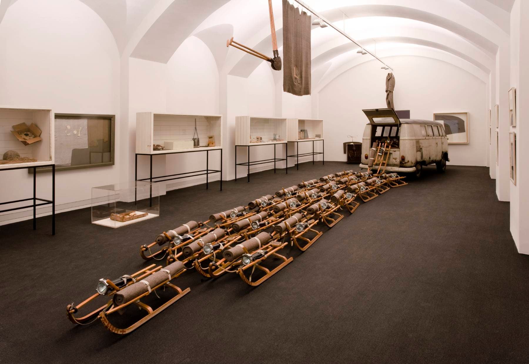 Kunstinstallation mit einem Oldtimer-Van, der mit mehreren Holzschlitten verbunden ist, in einer Galerie mit weißen Wänden und gewölbten Decken.