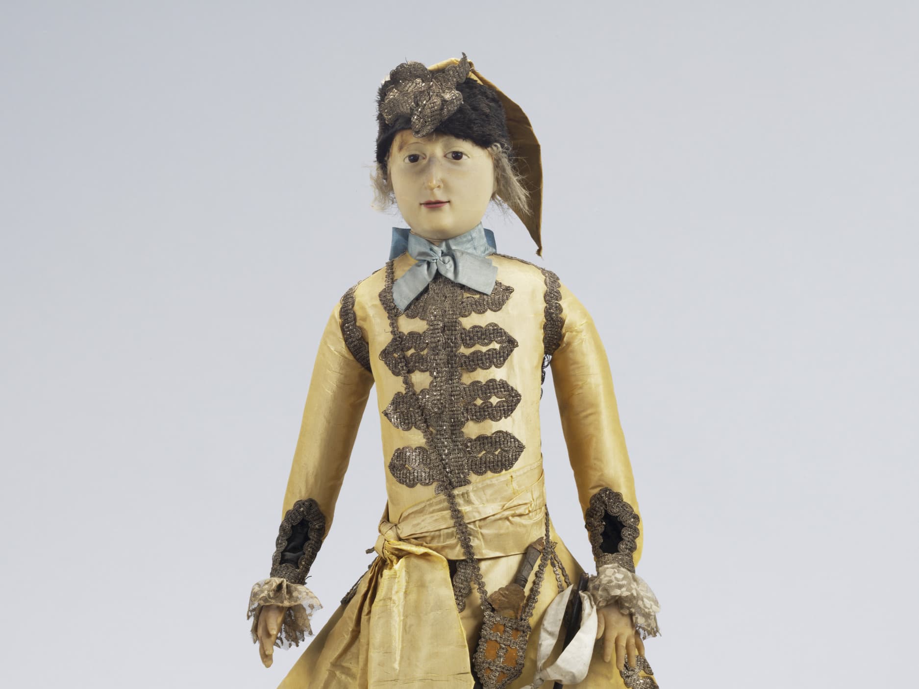 Antike Puppe in einem historischen Kostüm mit komplizierten Mustern, die einen Hut trägt und eine kleine Tasche hält, vor einem schlichten Hintergrund.