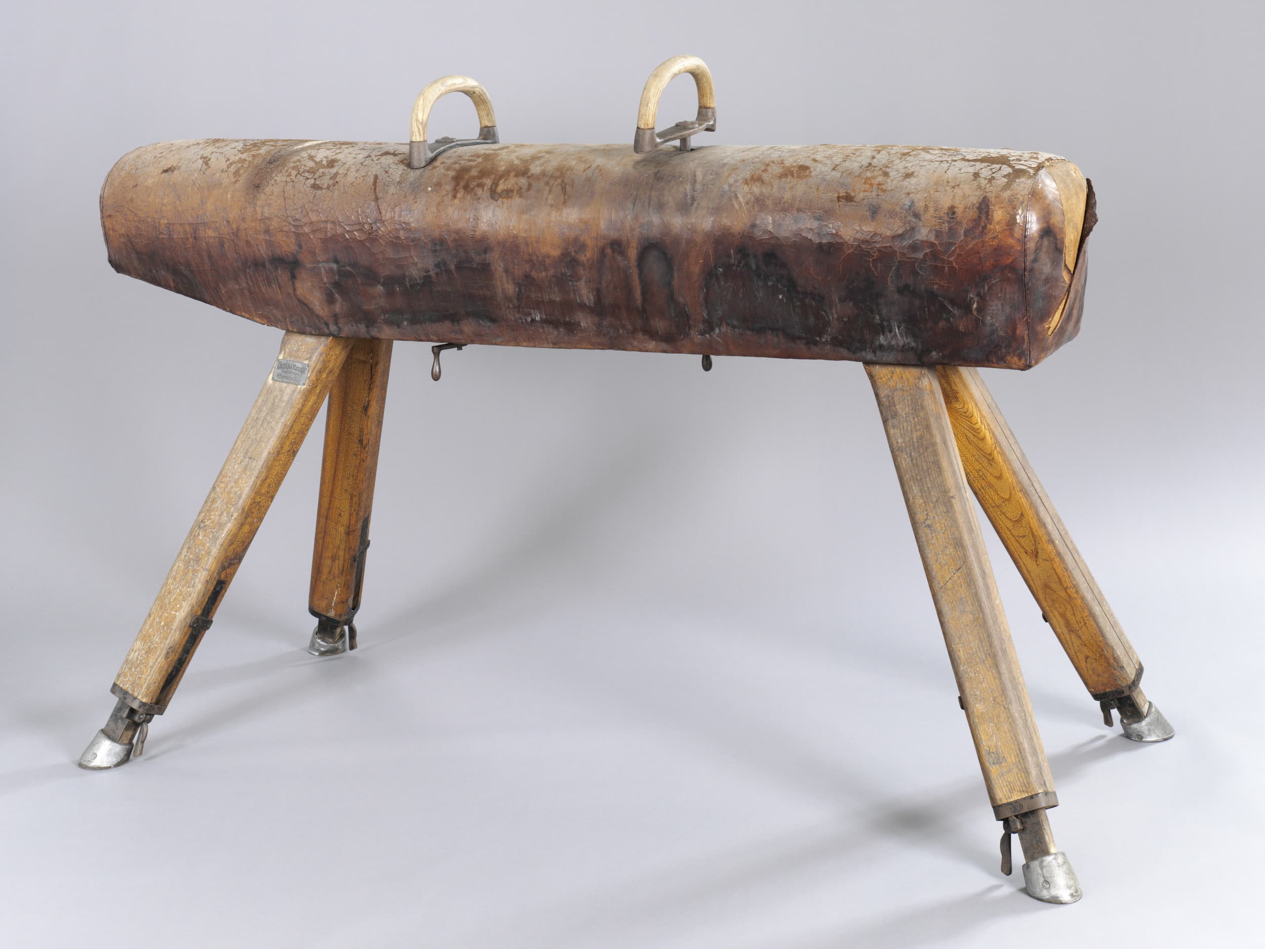 Vintage-Gymnastik-Pauschenpferd mit abgenutzter Lederoberseite, zwei Metallgriffen und Holzbeinen auf grauem Hintergrund.