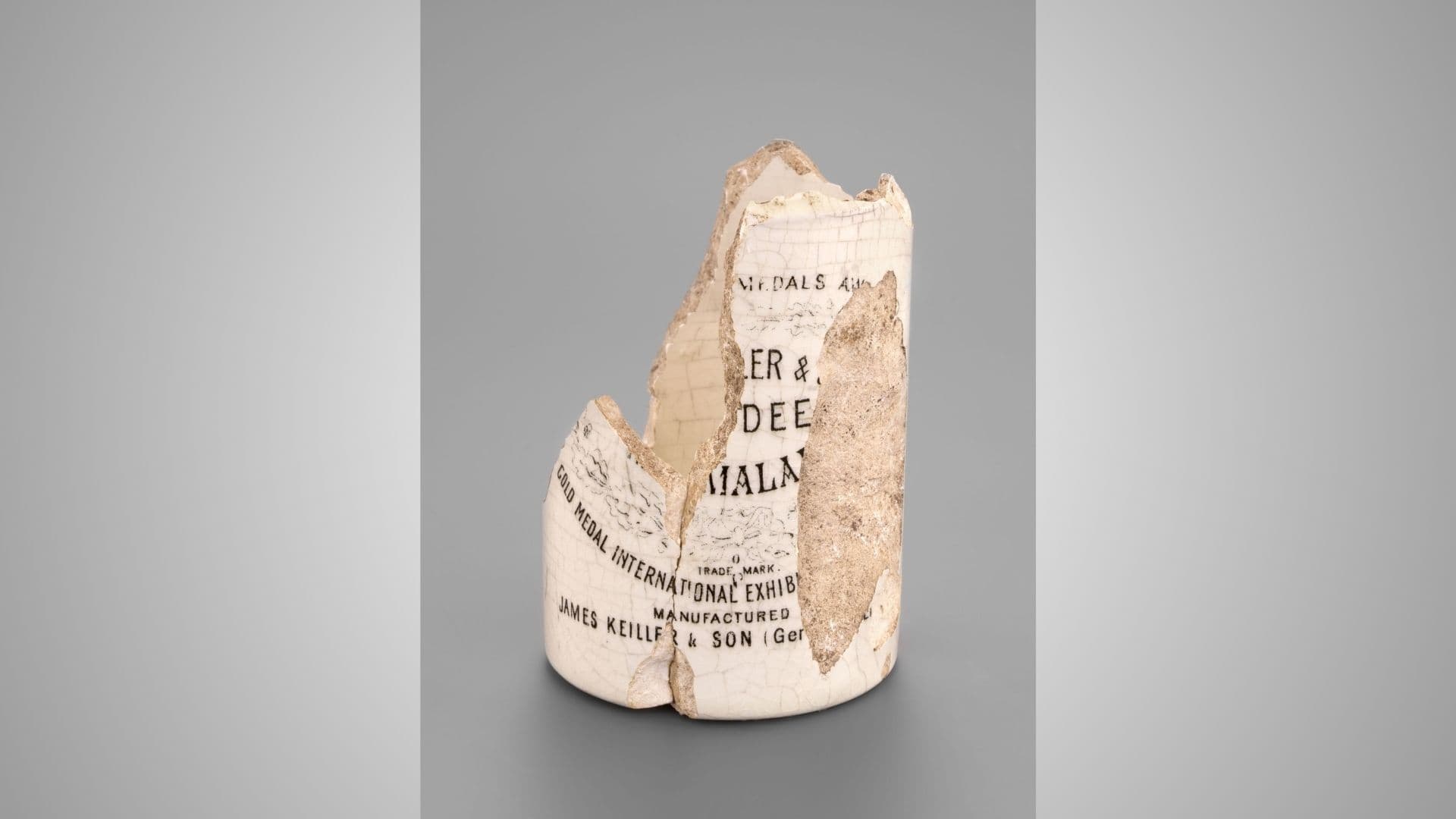 A broken ceramic jar with visible text and labels, featuring at aged and weathered appearance, set against a plain gray background.