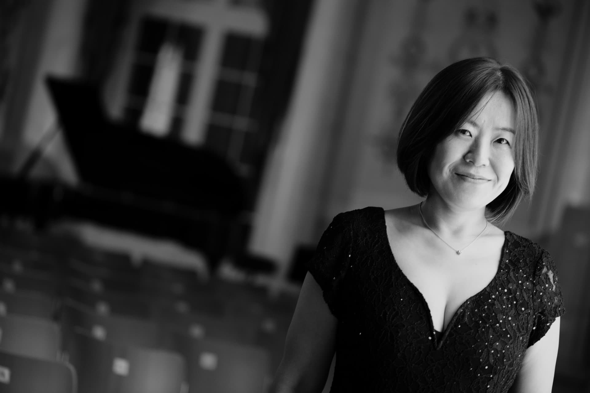 A woman in a black dress stands in a concert hall with a grand piano and rows of empty seats in the background, smiling slightly.
